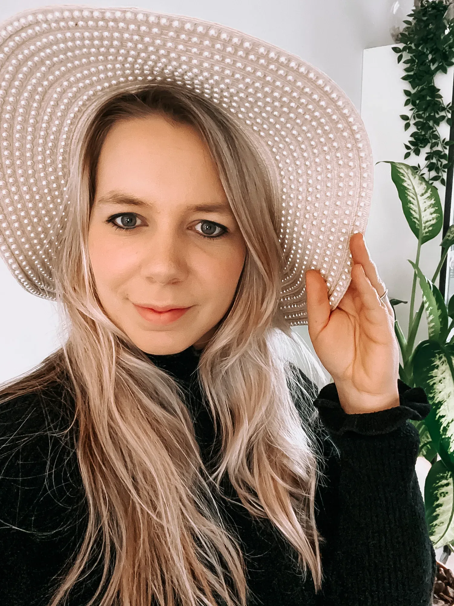 Eine Frau mit langen, blonden Haaren trägt einen selbstgemachten Strohhut, der mit weißen Perlen verziert ist. Sie lächelt in die Kamera und hält den Hut an der Krempe fest.