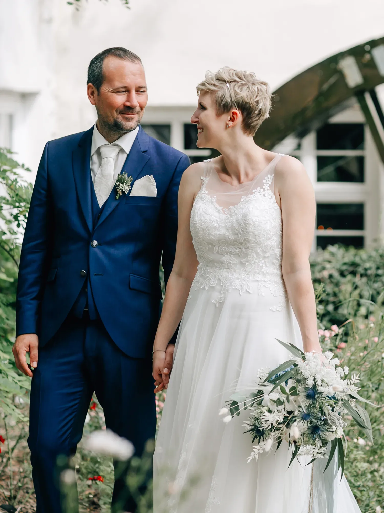 Ein Brautpaar spaziert Hand in Hand durch einen blühenden Garten. Der Bräutigam trägt einen dunkelblauen Anzug mit einer weißen Krawatte, während die Braut ein elegantes weißes Kleid mit floralen Details und einem Blumenstrauß aus weißen und blauen Blumen hält. Beide lächeln sich verliebt an.