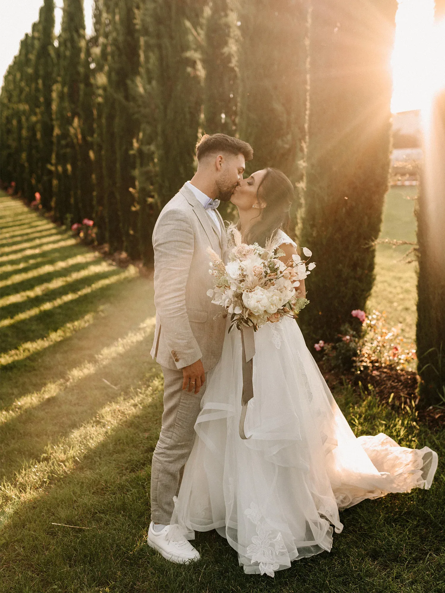 Ein Brautpaar steht bei Sonnenuntergang in einem Garten zwischen hohen Zypressen. Die Braut trägt ein weißes, romantisches Kleid und hält einen üppigen Blumenstrauß, während sie ihren Bräutigam, in einem hellen Anzug, liebevoll küsst.