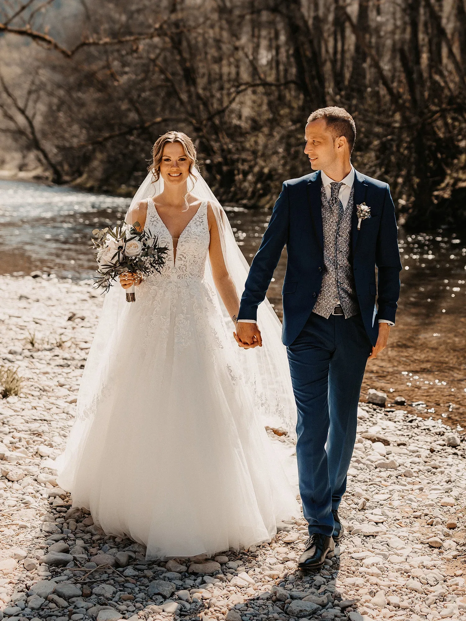 Ein Brautpaar spaziert Hand in Hand an einem sonnigen Tag entlang eines Flusses, die Braut trägt ein elegantes weißes Spitzenkleid und der Bräutigam einen blauen Anzug.