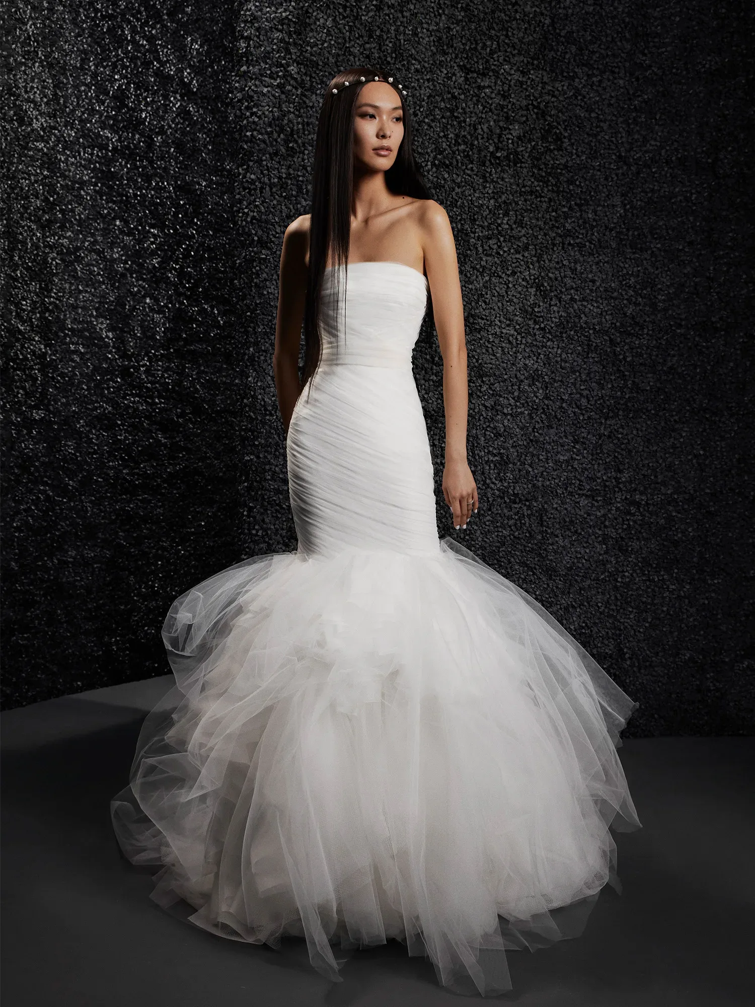 Eine Braut trägt ein schulterfreies, eng anliegendes Meerjungfrauen-Brautkleid aus Tüll mit einer voluminösen, gerafften Rockpartie. Das Kleid betont die schlanke Silhouette der Braut, während der dunkle Hintergrund die Eleganz des Designs hervorhebt.