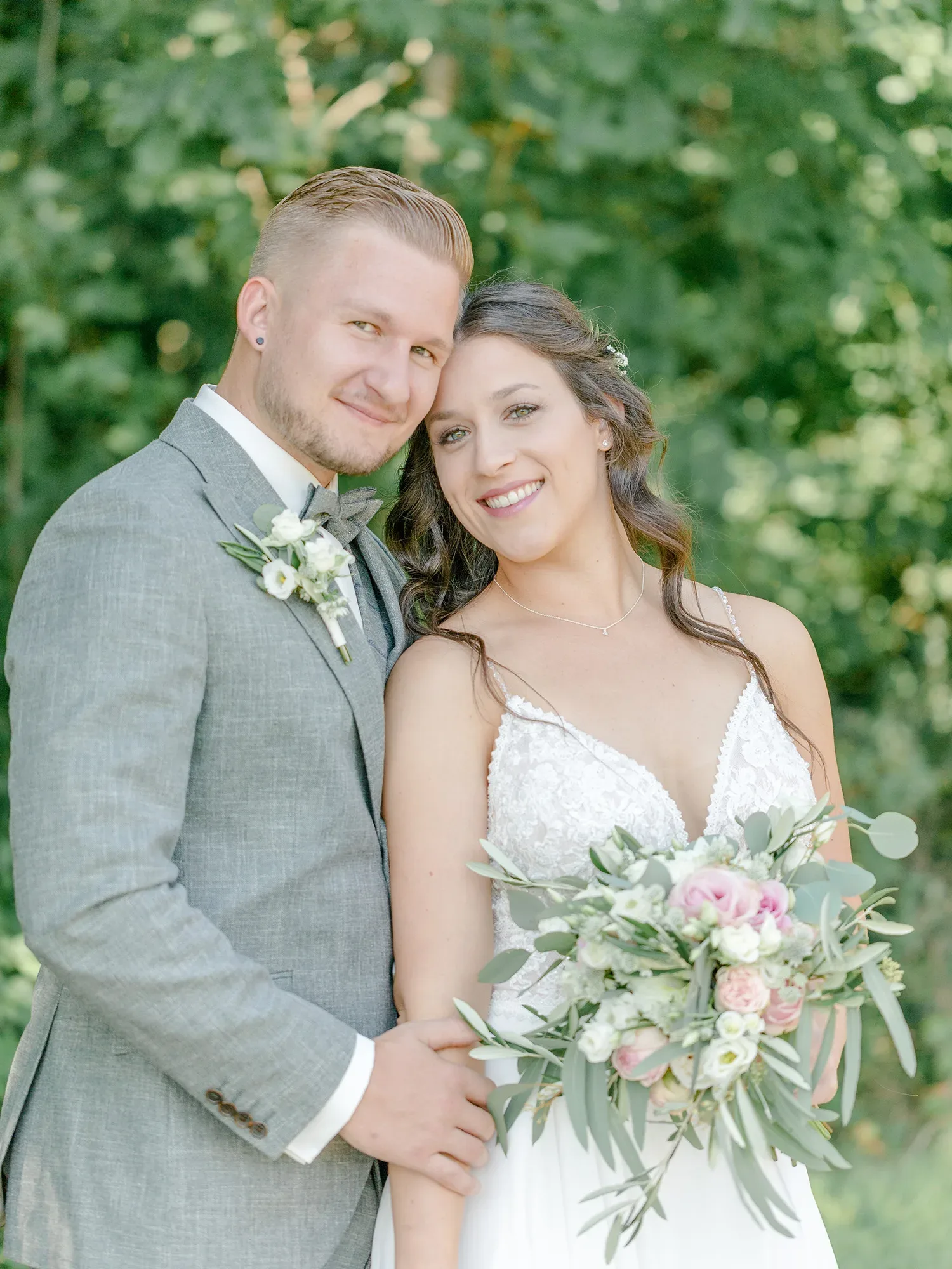 Ein lächelndes Brautpaar steht eng umschlungen im Freien, umgeben von grünem Laub. Der Bräutigam trägt einen grauen Anzug und die Braut ein Spitzenkleid mit einem zarten Blumenstrauß aus weißen und rosa Blumen.