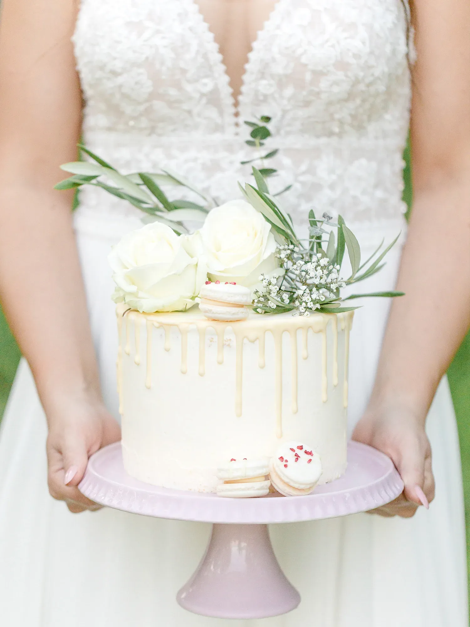 Eine Braut hält eine weiße Hochzeitstorte auf einer rosa Tortenplatte. Die Torte ist mit tropfender Glasur verziert und mit weißen Rosen, Macarons und Grün dekoriert. Im Hintergrund ist das Spitzenoberteil ihres Brautkleides sichtbar.