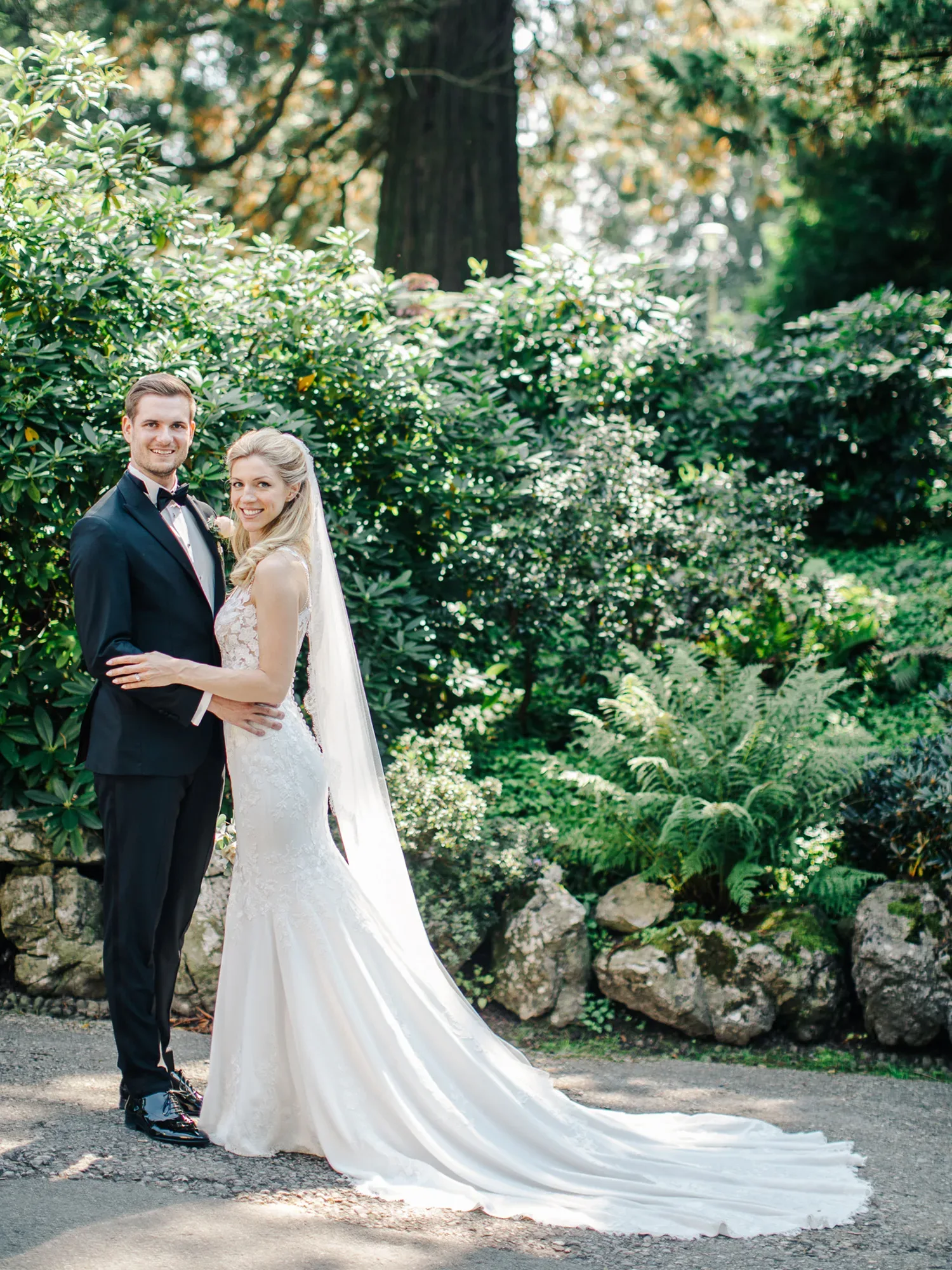 Das Brautpaar steht Arm in Arm in einem üppig grünen Garten. Die Braut trägt ein elegantes, weißes Kleid mit langer Schleppe, während der Bräutigam einen schwarzen Anzug und eine Fliege trägt. Beide lächeln glücklich in die Kamera.