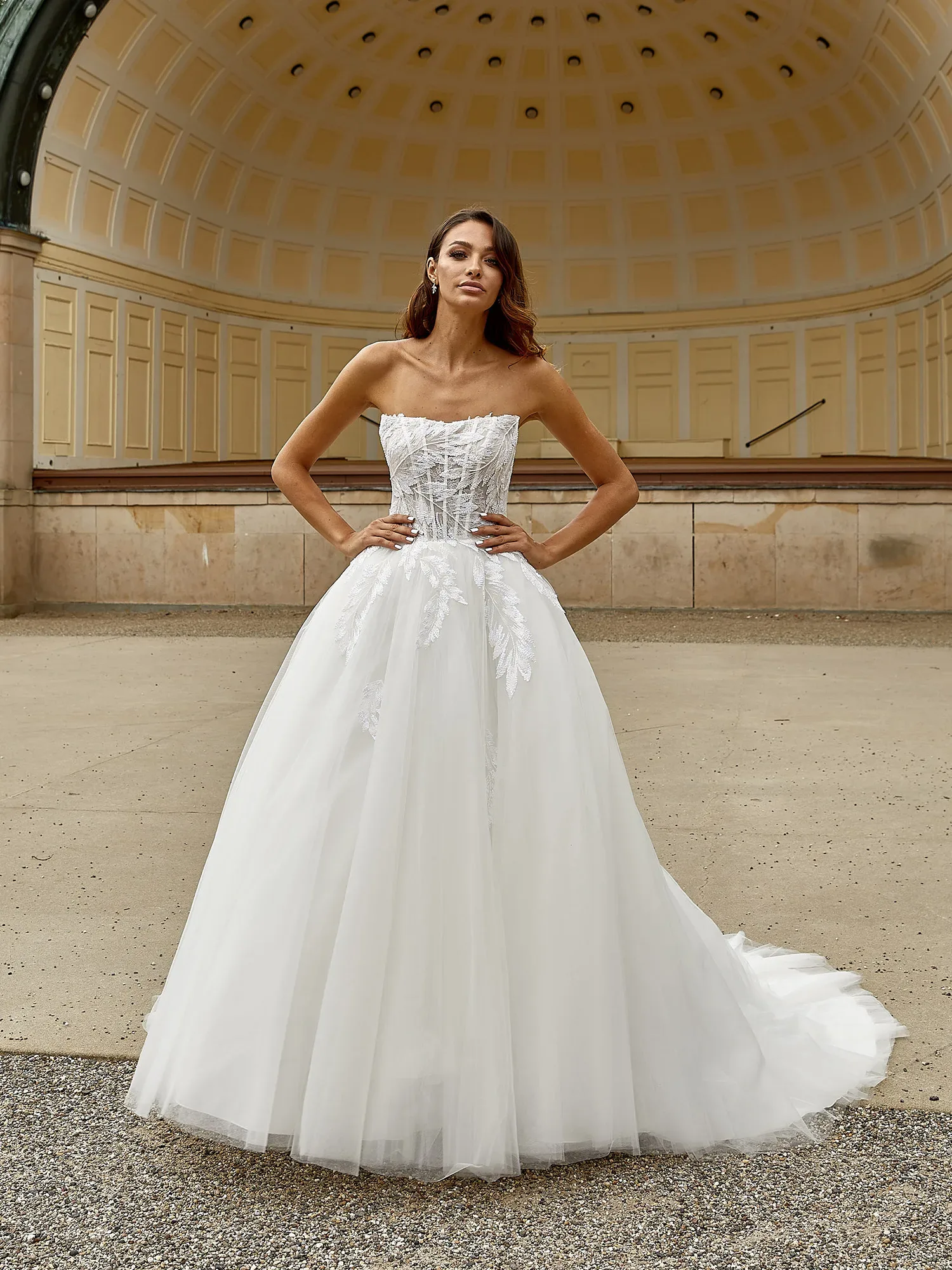 Eine Braut posiert in einem schulterfreien, detailreichen Ballkleid mit einem bestickten Oberteil und voluminösem Tüllrock vor einem historischen Pavillon. Die elegante Kulisse betont den klassischen Look des Brautkleides.