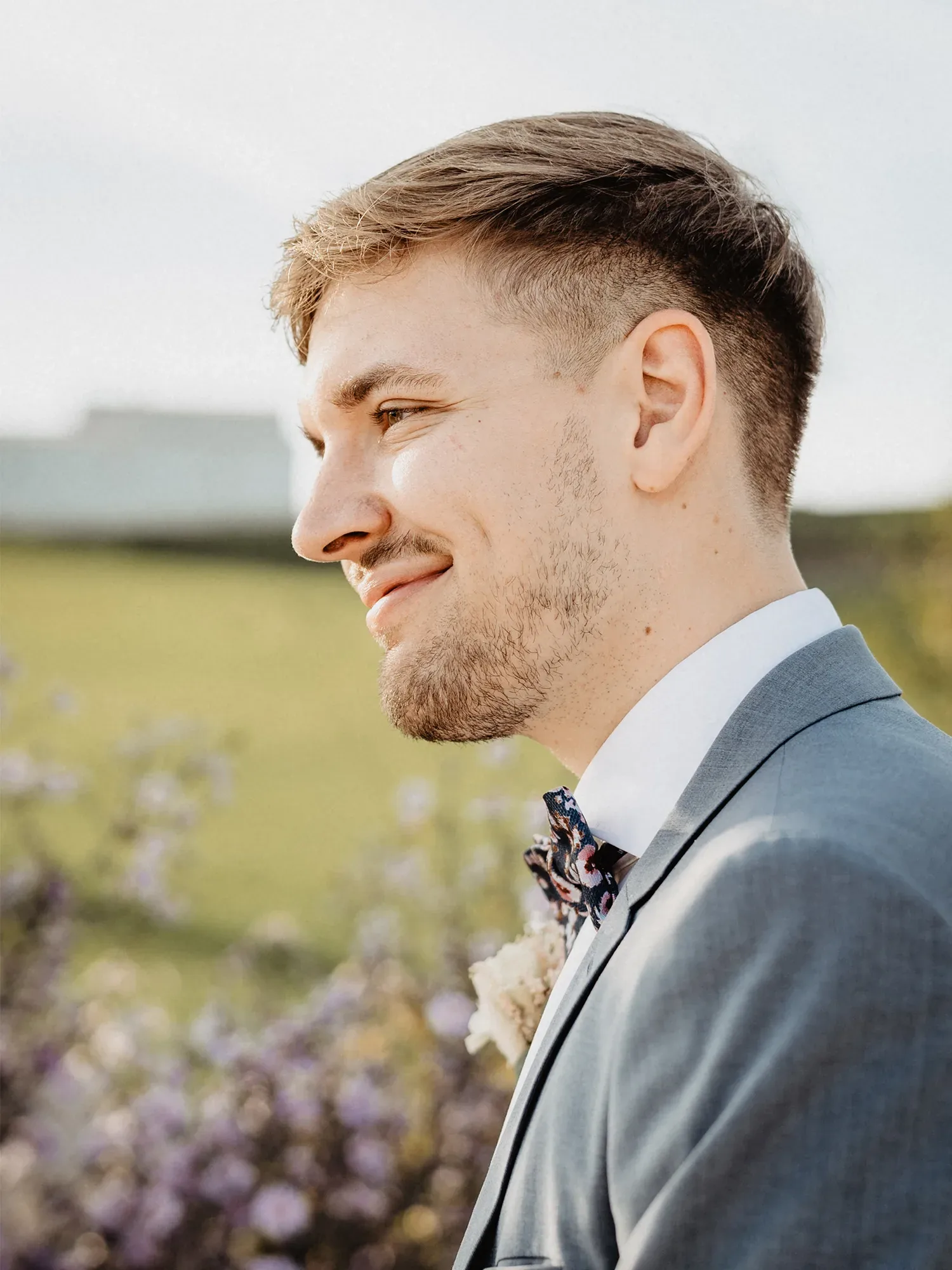 Ein junger Mann im Profil trägt einen eleganten grauen Anzug und eine gemusterte Fliege. Er lächelt leicht, während er vor einem blühenden, ländlichen Hintergrund steht.