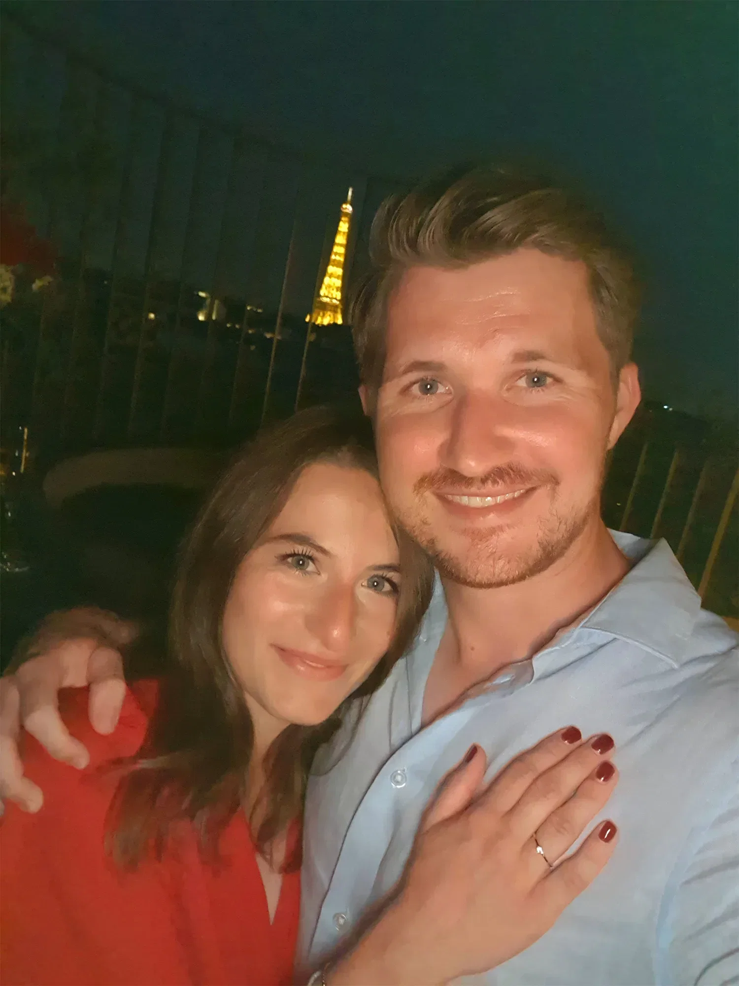 Ein Paar posiert lächelnd für ein Selfie vor dem Eiffelturm bei Nacht. Die Frau zeigt stolz ihren Verlobungsring, während sie den Mann umarmt.