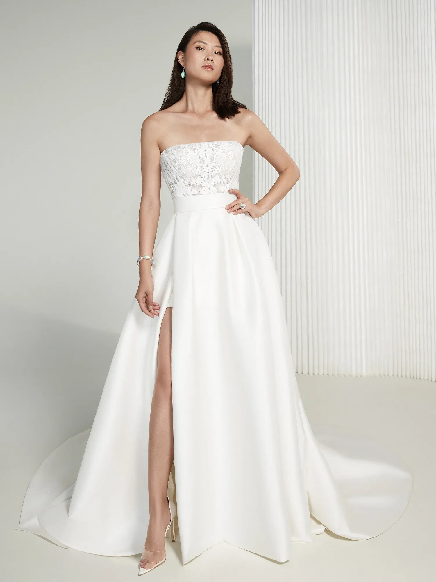 Eine Braut in einem eleganten, trägerlosen Brautkleid mit Spitzenoberteil und hohem Beinschlitz. Das fließende, bodenlange Kleid kombiniert klassische Eleganz mit modernen Details.
