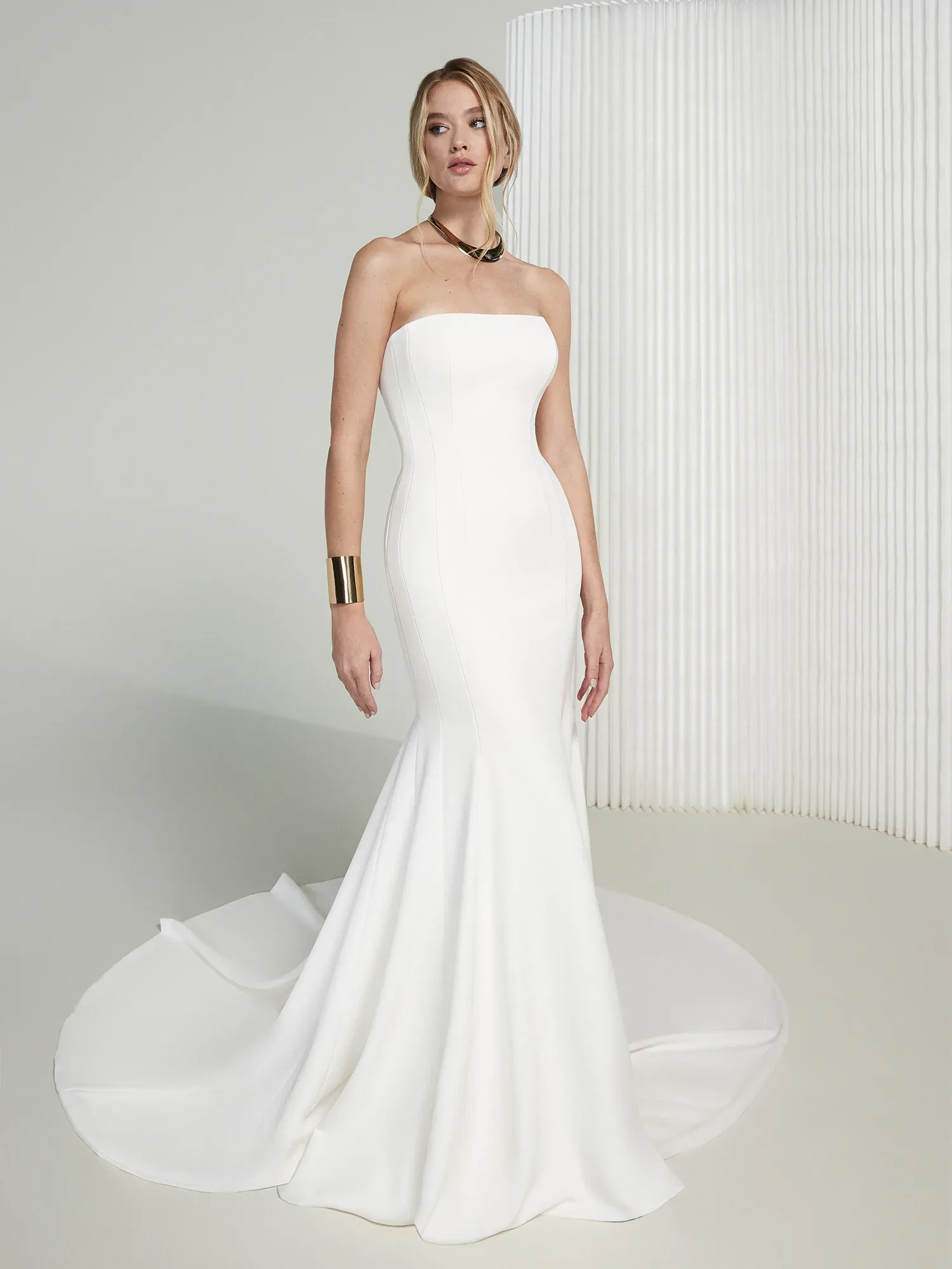 Eine Braut trägt ein minimalistisches, schulterfreies Meerjungfrauen-Brautkleid aus glattem Stoff. Das Kleid betont ihre schlanke Figur und endet in einer eleganten, schlichten Schleppe. Sie trägt ein goldfarbenes Armband und eine Halskette, die das moderne, stilvolle Design ergänzen.