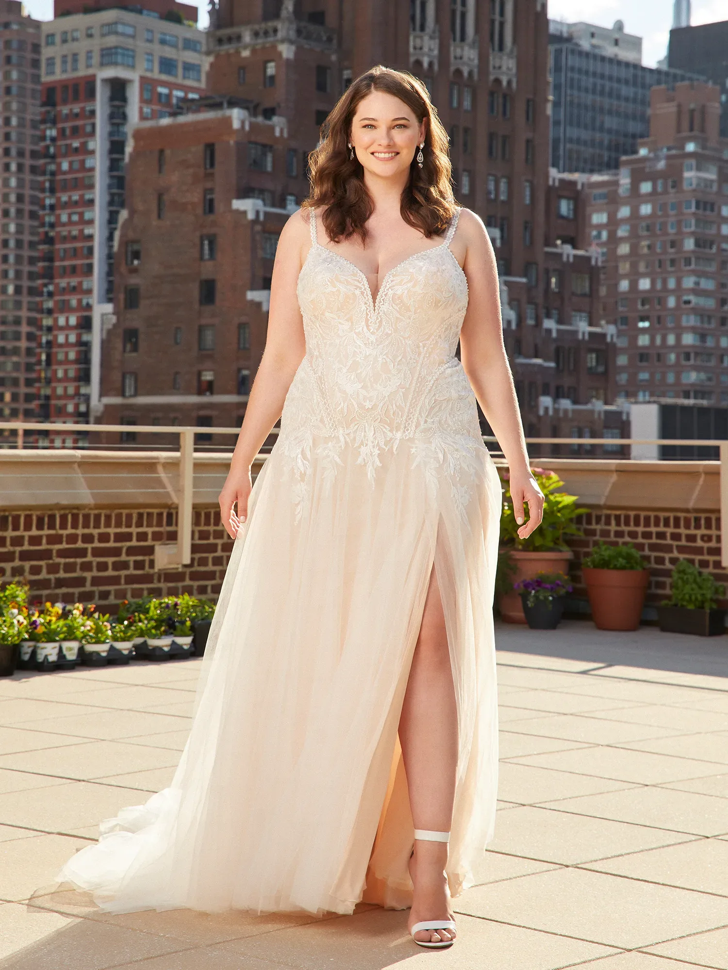 Eine lächelnde Frau in einem ärmellosen Brautkleid mit tiefem Ausschnitt und einer hohen Beinschlitze geht auf einer sonnigen Dachterrasse. Im Hintergrund sind hohe, städtische Gebäude zu sehen.