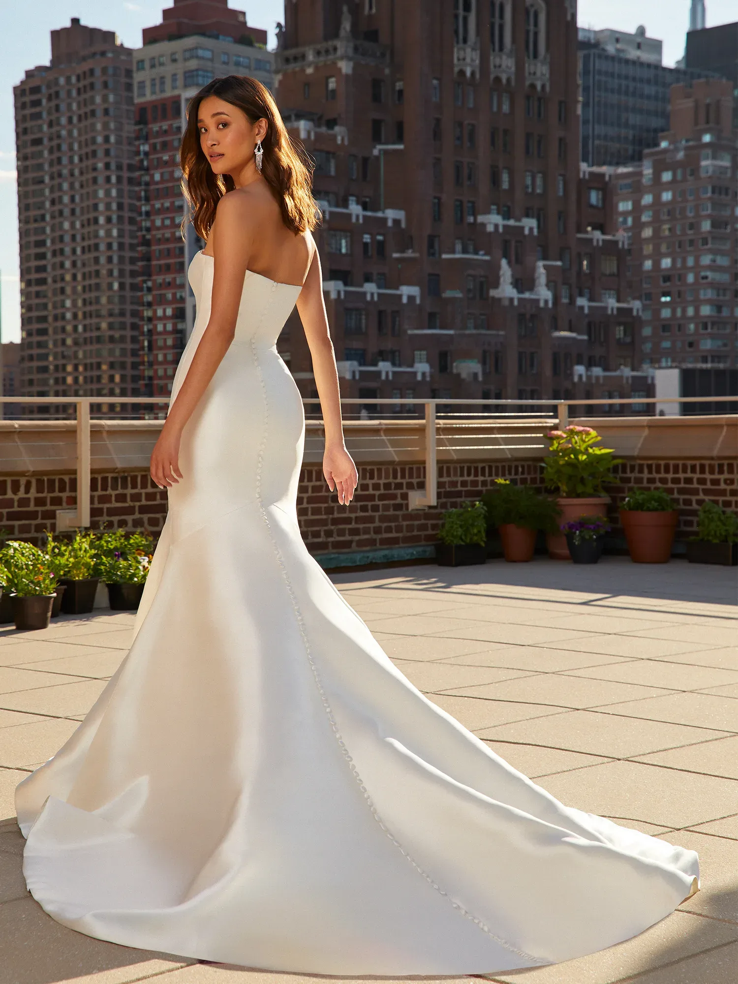 Eine Frau in einem eleganten, schulterfreien Brautkleid steht auf einer sonnigen Dachterrasse, die von modernen Gebäuden umgeben ist. Das Kleid hat eine lange Schleppe, die über den Boden gleitet.