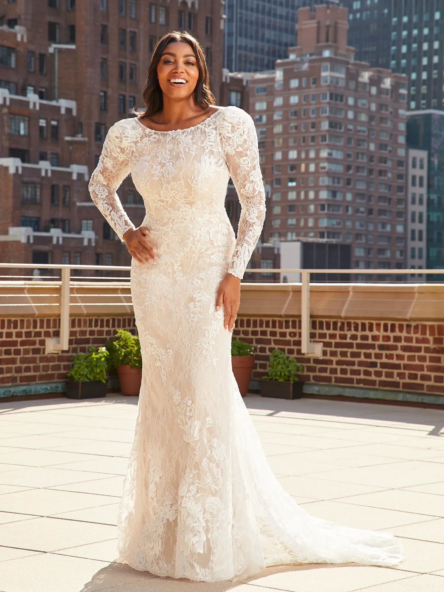 Eine Frau steht auf einer sonnigen Dachterrasse, gekleidet in ein elegantes, langärmeliges Spitzen-Brautkleid mit floralen Details. Sie lächelt und blickt in die Kamera, während im Hintergrund moderne Hochhäuser zu sehen sind.