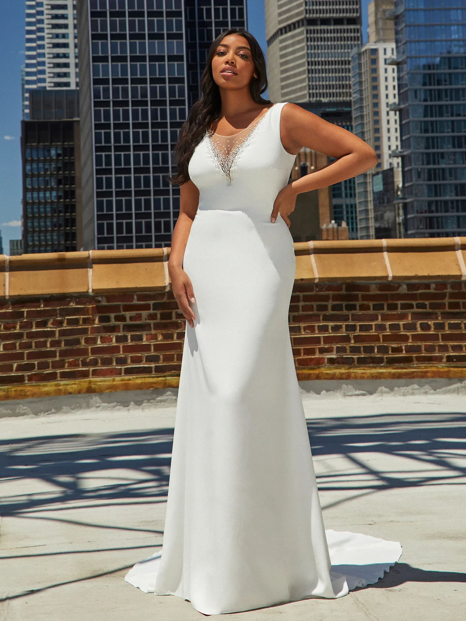 Eine Frau steht selbstbewusst in einem schlichten, ärmellosen weißen Brautkleid auf einer urbanen Dachterrasse. Hinter ihr ragen moderne Wolkenkratzer in den Himmel, die den Kontrast zwischen moderner Architektur und klassischer Brautmode betonen.