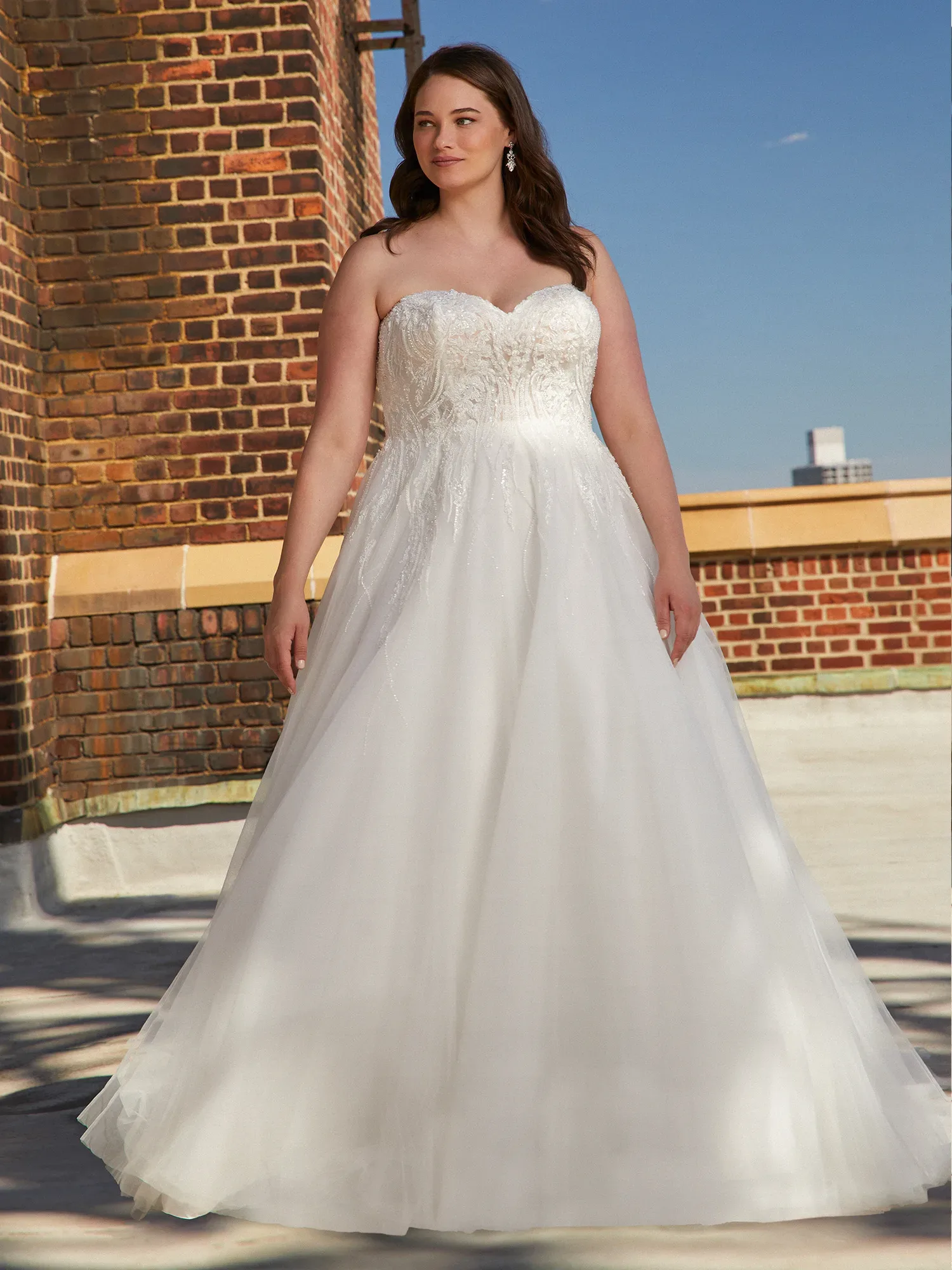 Eine Frau trägt ein trägerloses, klassisches Brautkleid mit A-Linien-Rock. Das Kleid besticht durch zarte Stickereien auf dem Mieder und einen fließenden Rock, während sie auf einer Terrasse vor einer Backsteinmauer posiert.
