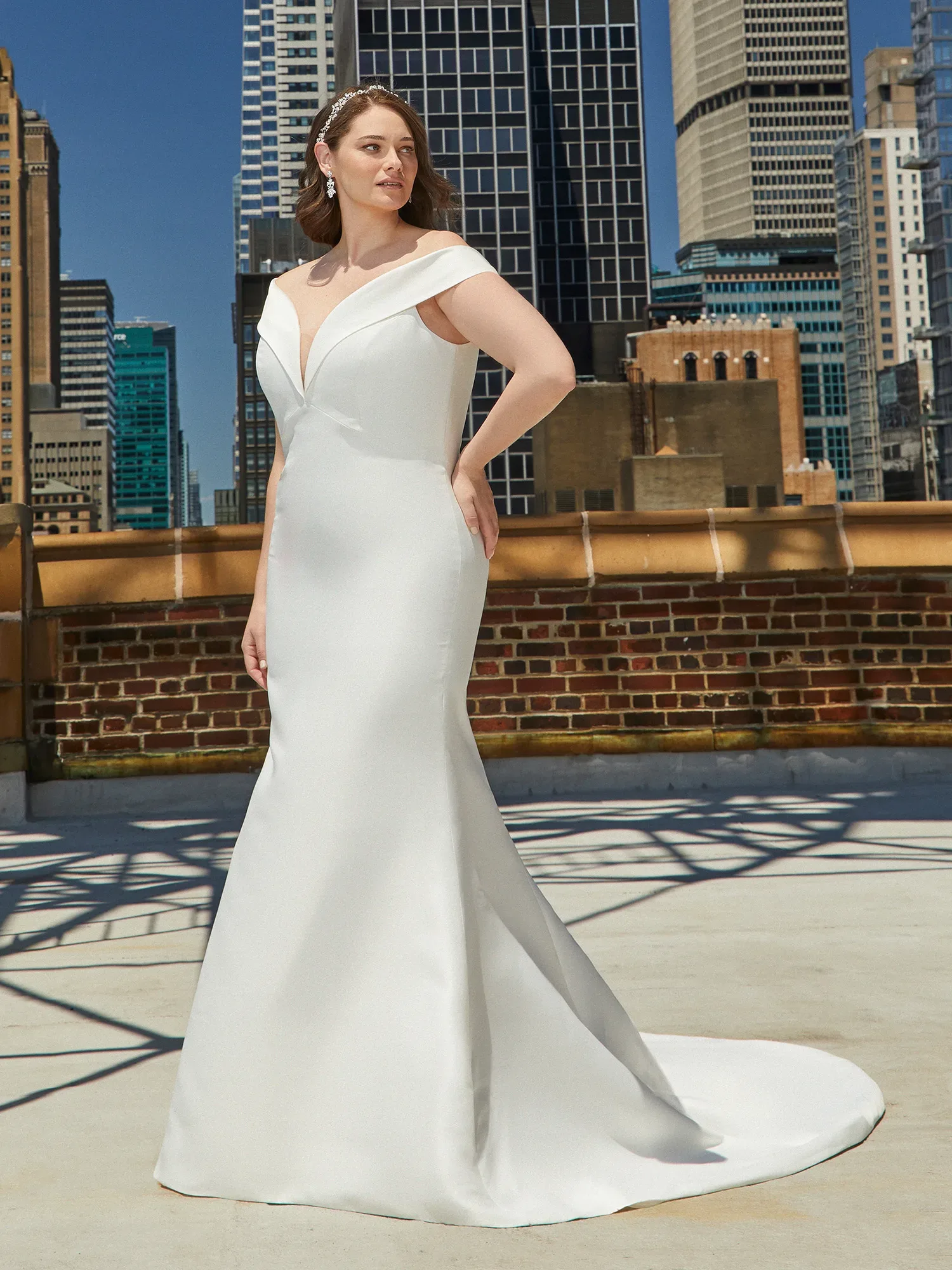 Eine Frau trägt ein schlichtes, aber elegantes Brautkleid mit tiefem V-Ausschnitt und breiten Trägern, die ihre Schultern umrahmen. Sie steht auf einer urbanen Dachterrasse vor einer Kulisse aus modernen Hochhäusern.