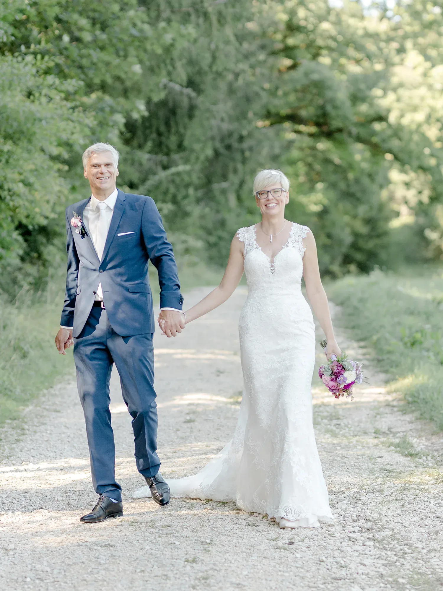 Sarah und Achim gehen Hand in Hand auf einem Kiesweg durch einen Wald. Die Braut trägt ein eng anliegendes, spitzenbesetztes weißes Hochzeitskleid und hält einen Blumenstrauß in Rosa- und Lilatönen. Der Bräutigam ist in einem eleganten blauen Anzug gekleidet. Beide lächeln und genießen den sonnigen Tag in der Natur.