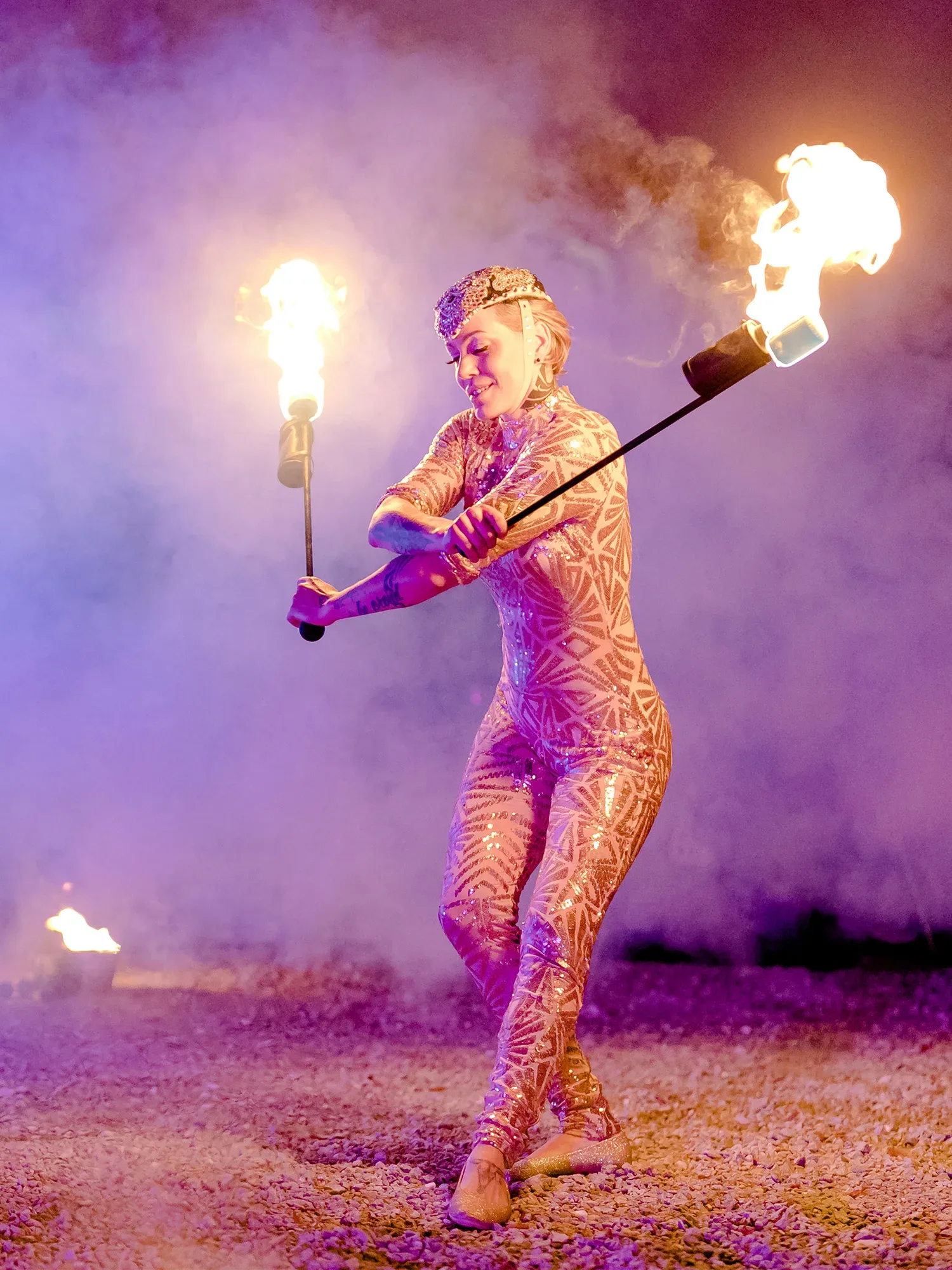 Eine Feuerkünstlerin führt eine atemberaubende Show mit brennenden Fackeln auf. Sie trägt einen glitzernden, hautengen Ganzkörperanzug mit geometrischen Mustern und bewegt die Fackeln geschickt durch die Luft. Im Hintergrund ist dicker Rauch zu sehen, der von den Flammen beleuchtet wird und eine dramatische Atmosphäre erzeugt.