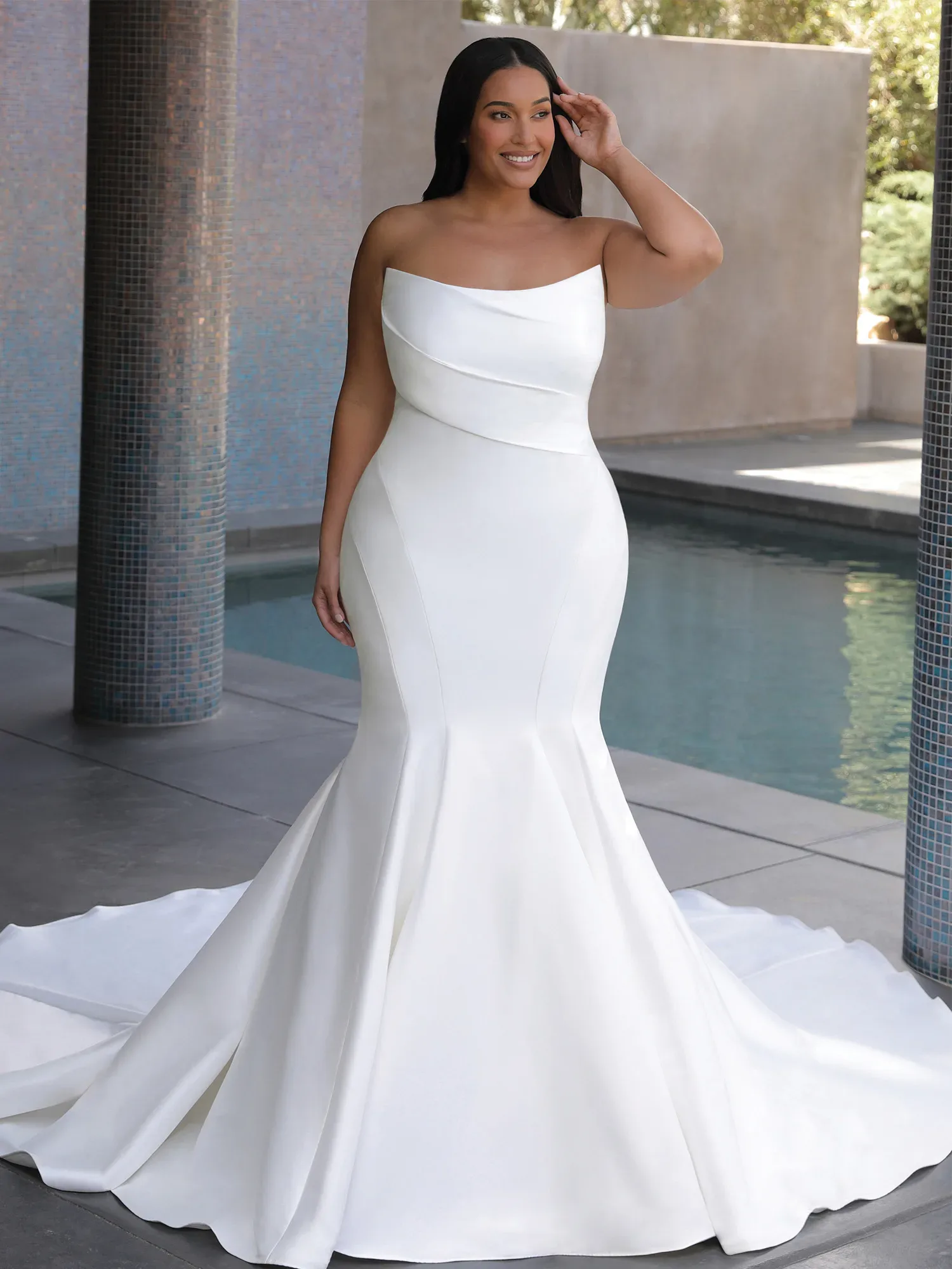 Eine Braut in einem modernen, schulterfreien Brautkleid aus Satin steht lächelnd neben einem Pool. Das figurbetonte Kleid betont ihre Kurven und endet in einem eleganten, bodenlangen Rock.