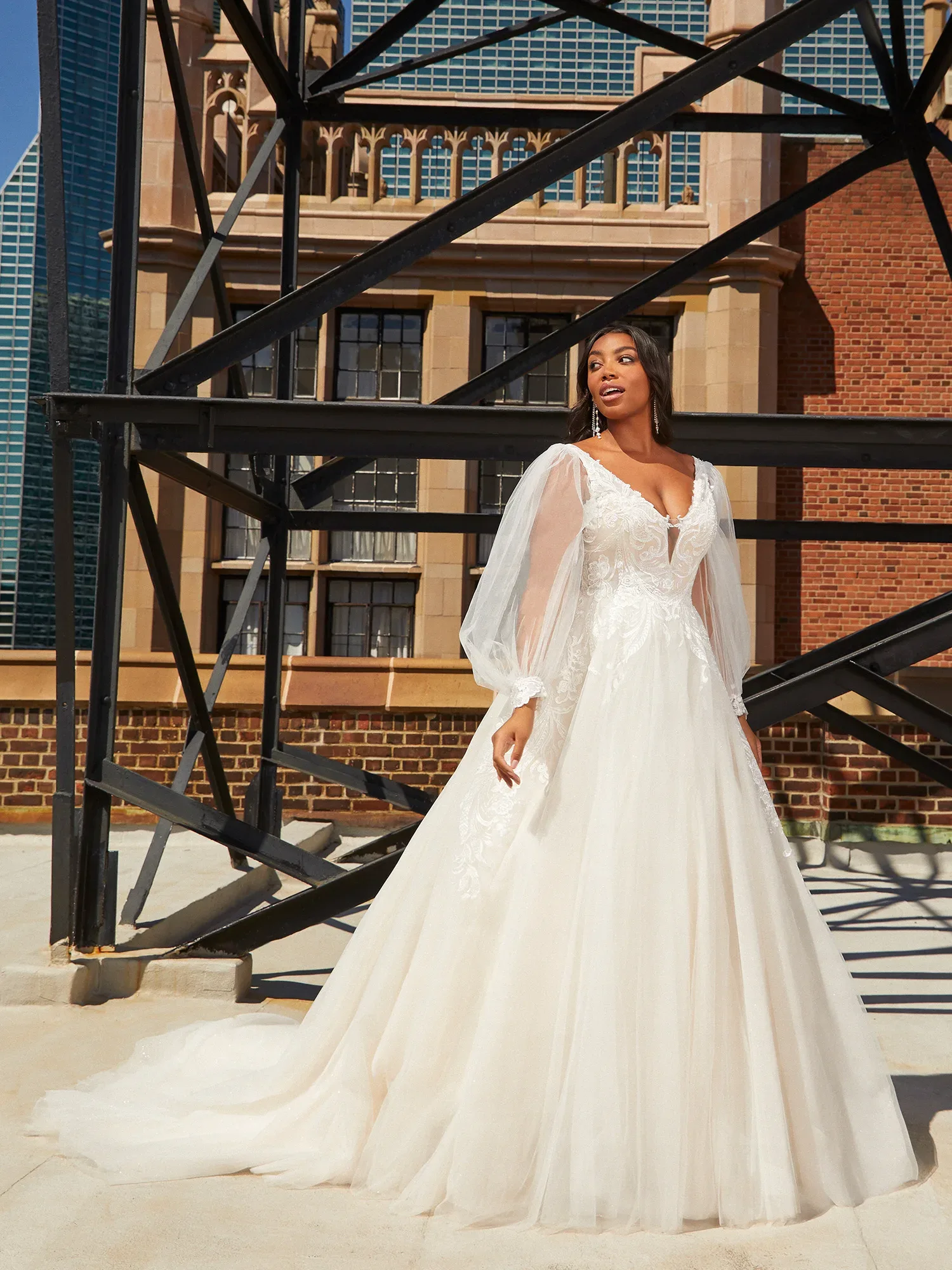 Eine Braut in einem voluminösen Spitzenkleid mit transparenten, langen Ärmeln und tiefem V-Ausschnitt steht auf einer Dachterrasse. Im Hintergrund sind Backsteingebäude und moderne Architektur zu sehen, die der Szene einen urbanen, industriellen Touch verleihen.