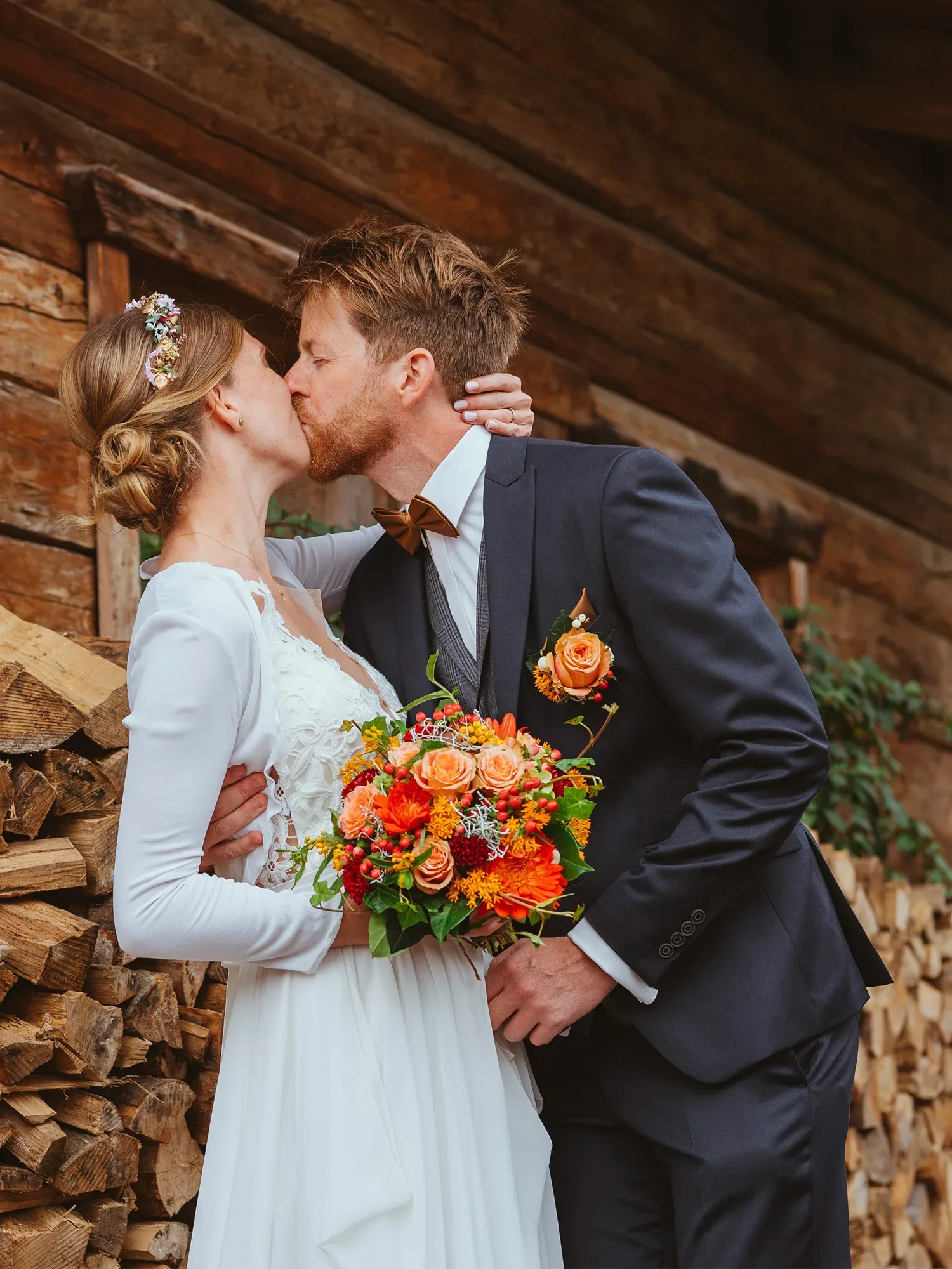 Ein Brautpaar, elegant gekleidet, steht vor einer rustikalen Holzwand und teilt einen Kuss. Die Braut trägt ein weißes Kleid und hält einen leuchtend orangefarbenen Blumenstrauß, der Bräutigam ist in einem dunklen Anzug gekleidet.