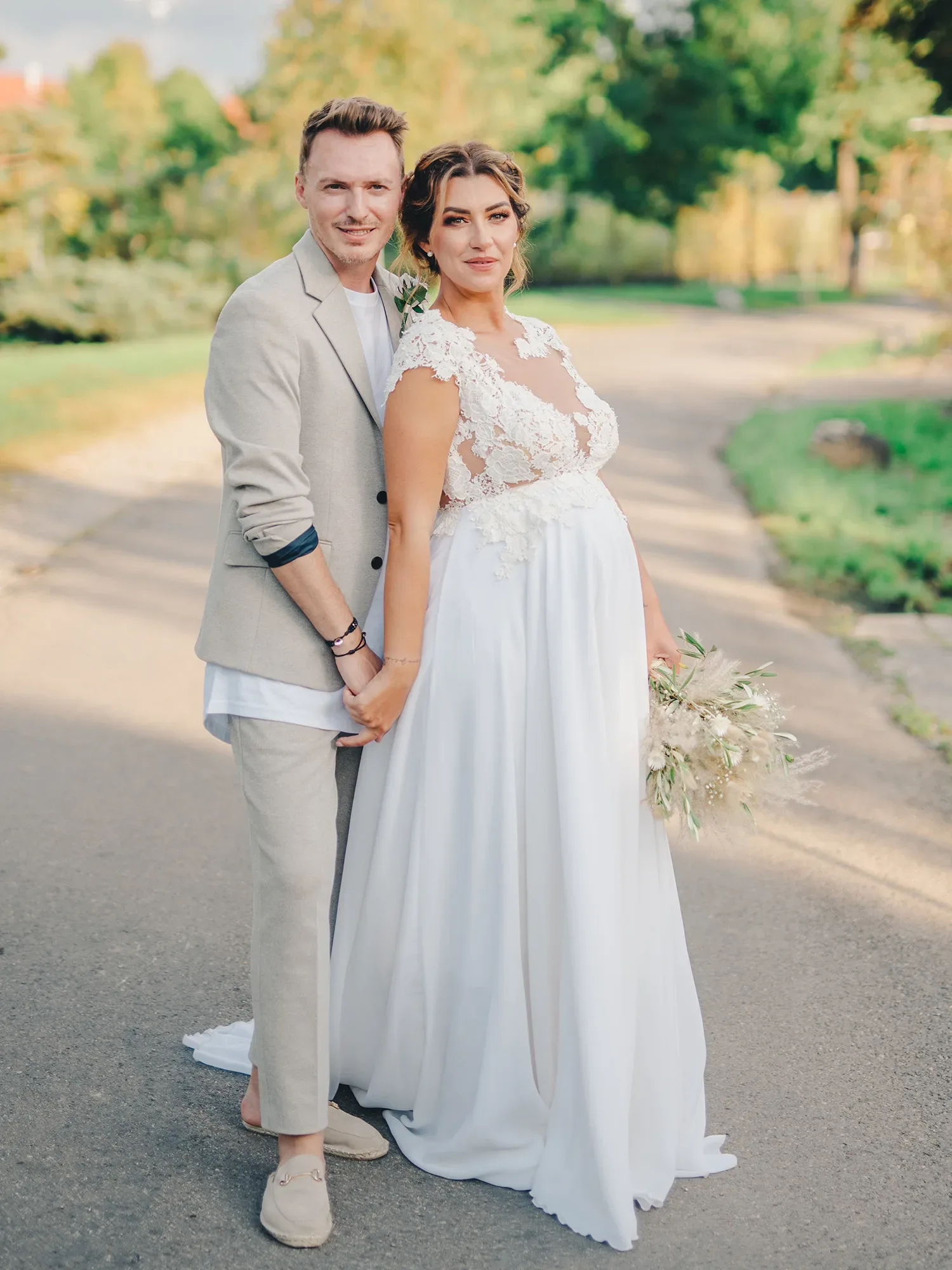 Das Brautpaar steht Hand in Hand auf einem Weg, umgeben von grüner Natur. Die Braut trägt ein weißes Kleid mit Spitzenoberteil, der Bräutigam einen hellen Anzug mit einem lockeren weißen Hemd. Beide blicken lächelnd in die Kamera.