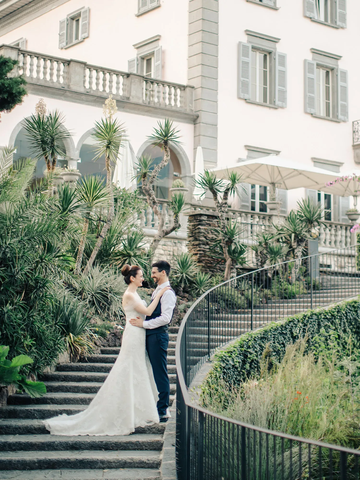 Das Brautpaar steht liebevoll umarmt auf einer Steintreppe vor einem mediterranen Herrenhaus. Die Braut trägt ein weißes Hochzeitskleid, und der Bräutigam ist in einen blauen Anzug mit Weste gekleidet. Umgeben von grünen Pflanzen strahlen sie Freude aus.
