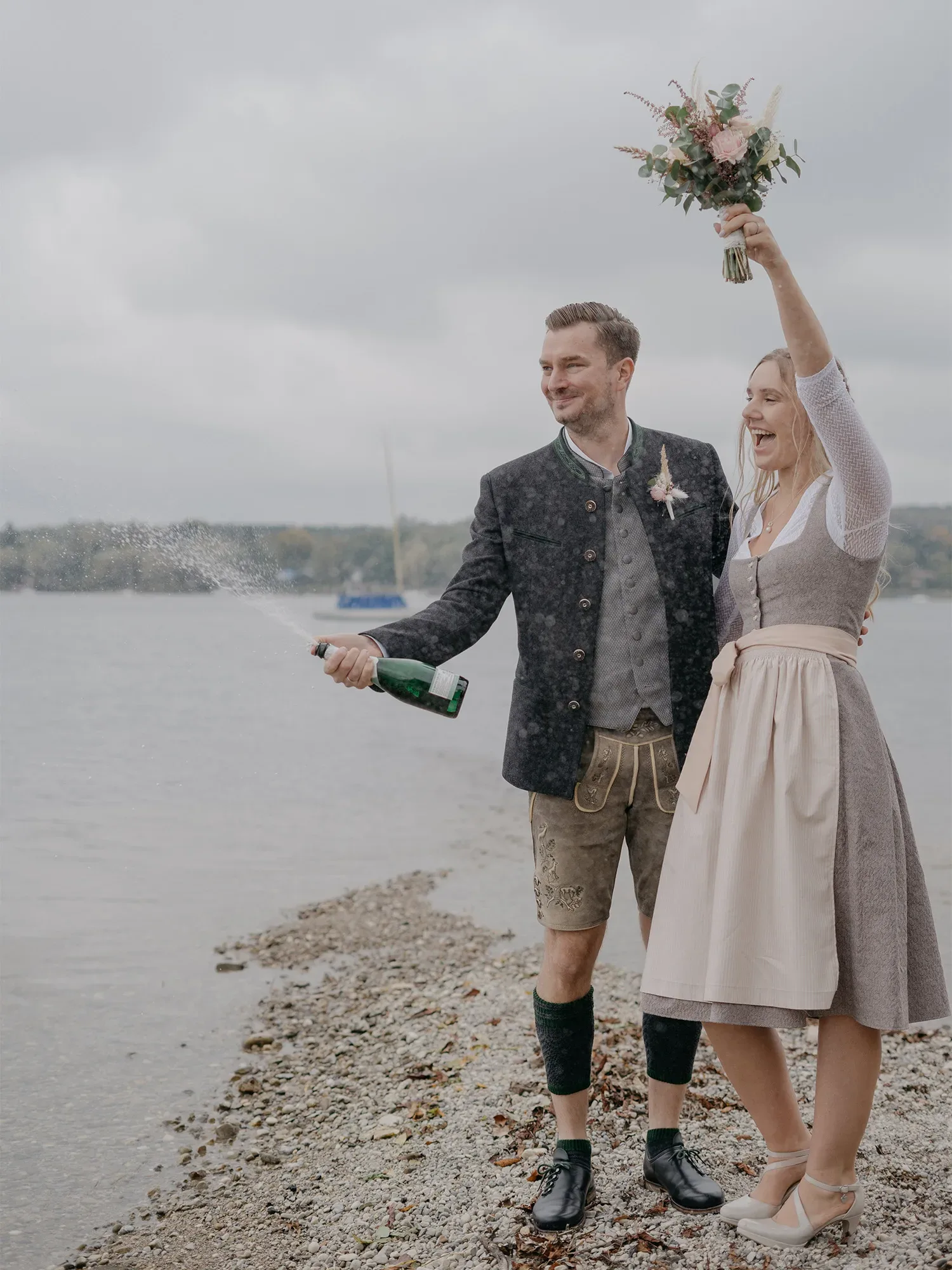 Ein Paar in bayerischer Tracht feiert am Ufer eines Sees. Der Bräutigam öffnet eine Sektflasche und sprüht Champagner, während die Braut ihren Arm mit einem Blumenstrauß in die Höhe streckt.