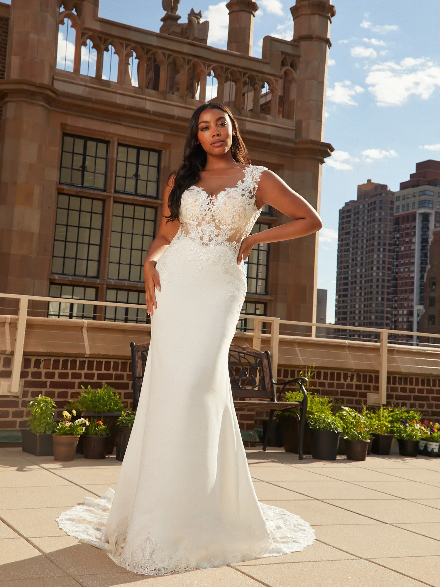 Eine Braut in einem figurbetonten weißen Kleid mit floralen Spitzenapplikationen und transparenten Details posiert auf einer Dachterrasse vor einem historischen Gebäude mit steinernen Bögen und Fenstern. Im Hintergrund ragen Hochhäuser in den Himmel.