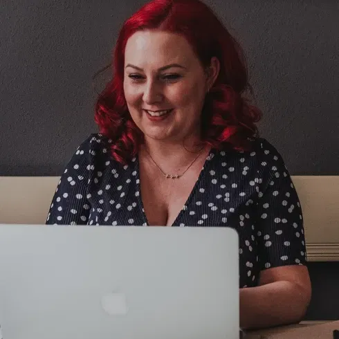 Frau mit roten Haaren lächelt, während sie an einem Laptop arbeitet. Sie trägt ein dunkelblaues, gepunktetes Kleid und sitzt in einem gemütlichen Büro.