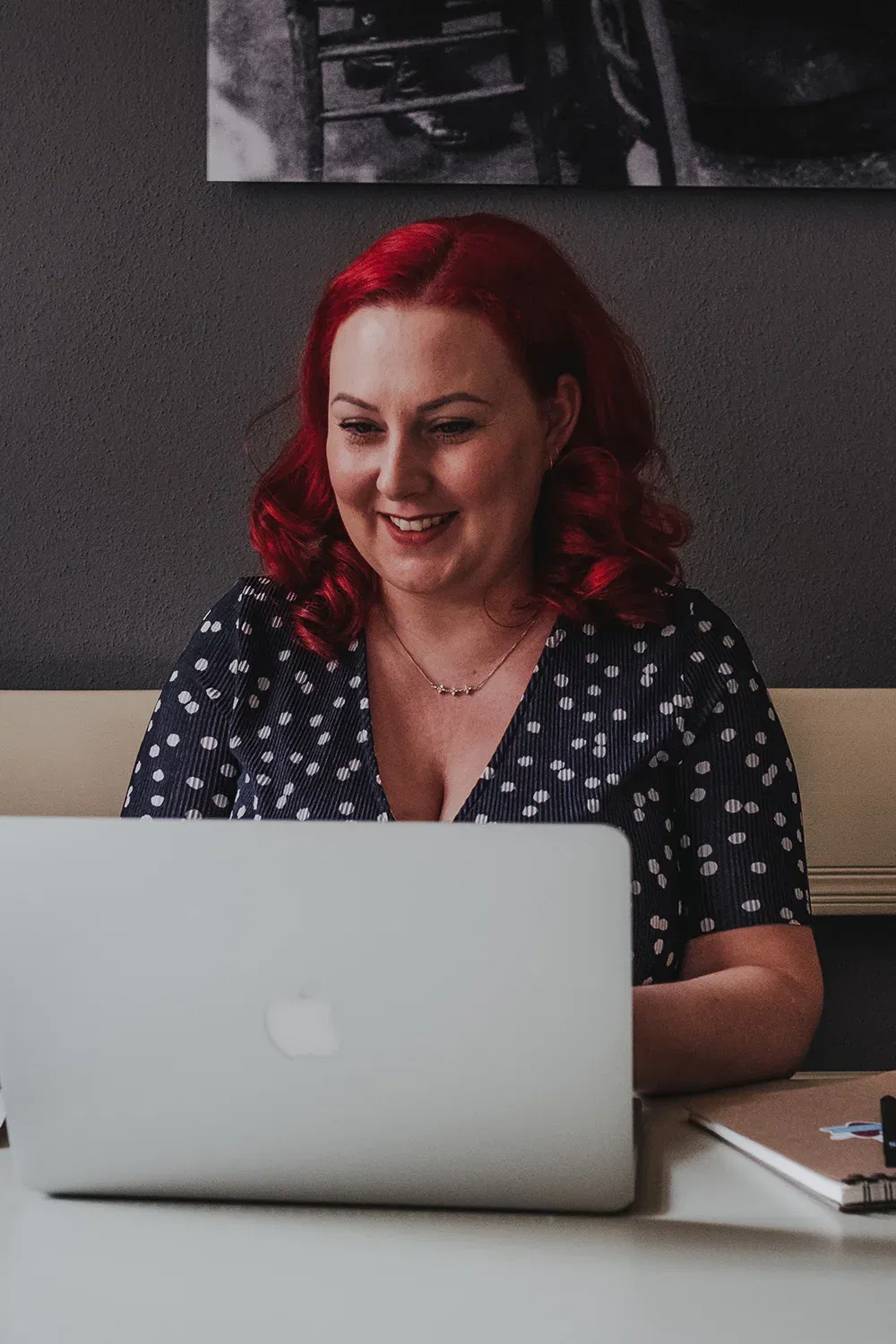 Frau mit roten Haaren lächelt, während sie an einem Laptop arbeitet. Sie trägt ein dunkelblaues, gepunktetes Kleid und sitzt in einem gemütlichen Büro.