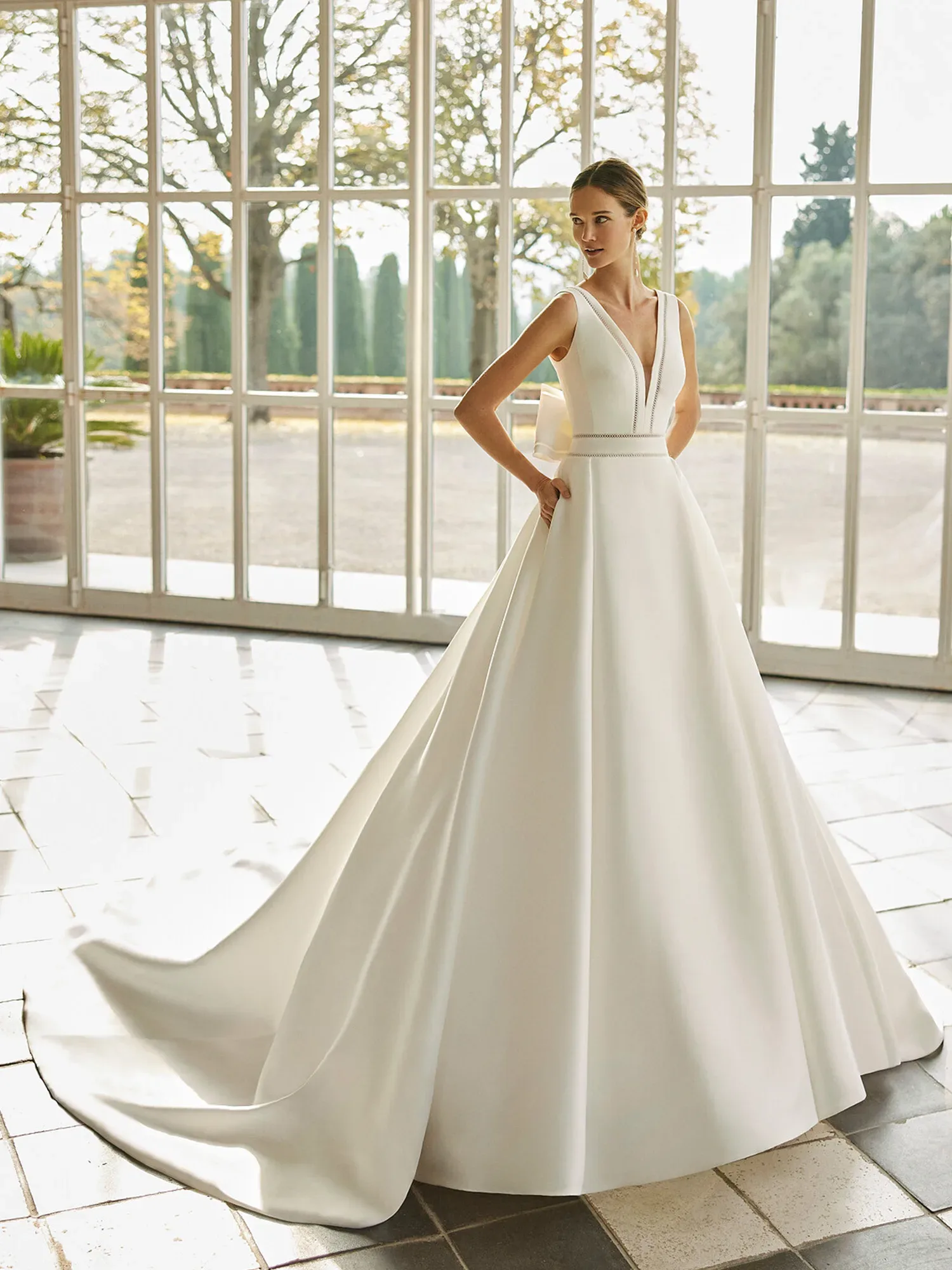 Eine Braut steht in einem lichtdurchfluteten Raum und trägt ein ärmelloses A-Linien-Hochzeitskleid mit tiefem V-Ausschnitt. Das Kleid hat eine lange Schleppe, und hinter ihr sind große Fenster, die auf eine Gartenlandschaft blicken.