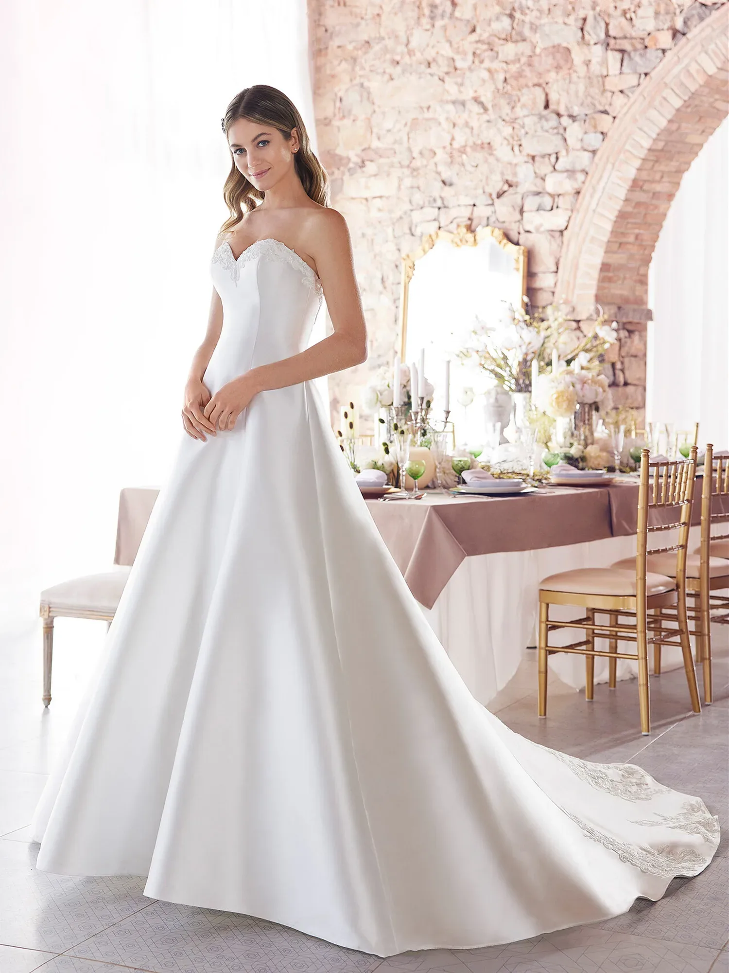 Eine Braut trägt ein klassisches trägerloses A-Linien-Hochzeitskleid mit Spitzenbesatz am Ausschnitt. Sie steht in einem eleganten Raum mit Steinwänden und einem gedeckten Tisch im Hintergrund und lächelt sanft.