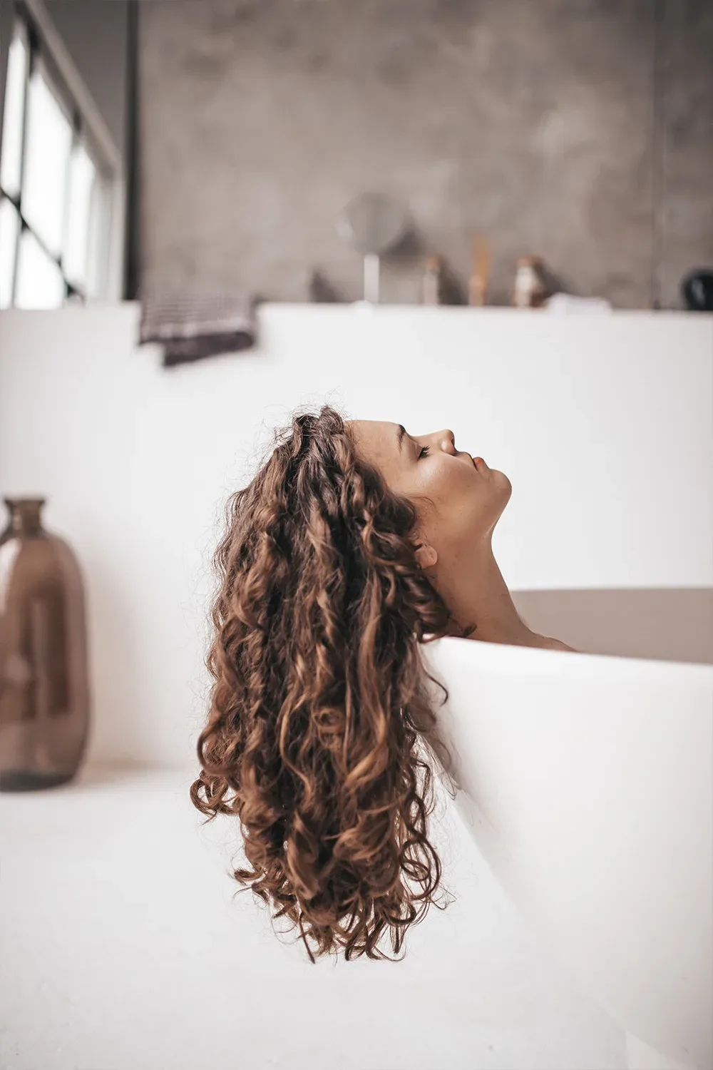 Eine Frau mit lockigem Haar lehnt entspannt in einer freistehenden Badewanne zurück. Ihr Haar hängt über den Rand der Wanne, während sie den Moment der Ruhe genießt. Die Szene vermittelt Entspannung in einem modernen Badezimmer.
