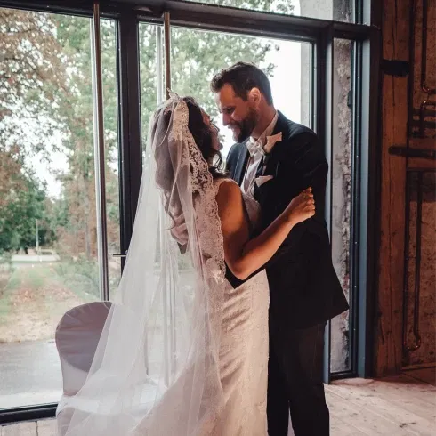 Ein Brautpaar steht in einem modernen, rustikalen Raum, während sie sich innig umarmen. Die Braut trägt ein langes, weißes Kleid mit Schleier, während der Bräutigam einen klassischen schwarzen Anzug trägt. Große Fenster im Hintergrund lassen natürliches Licht in den Raum.