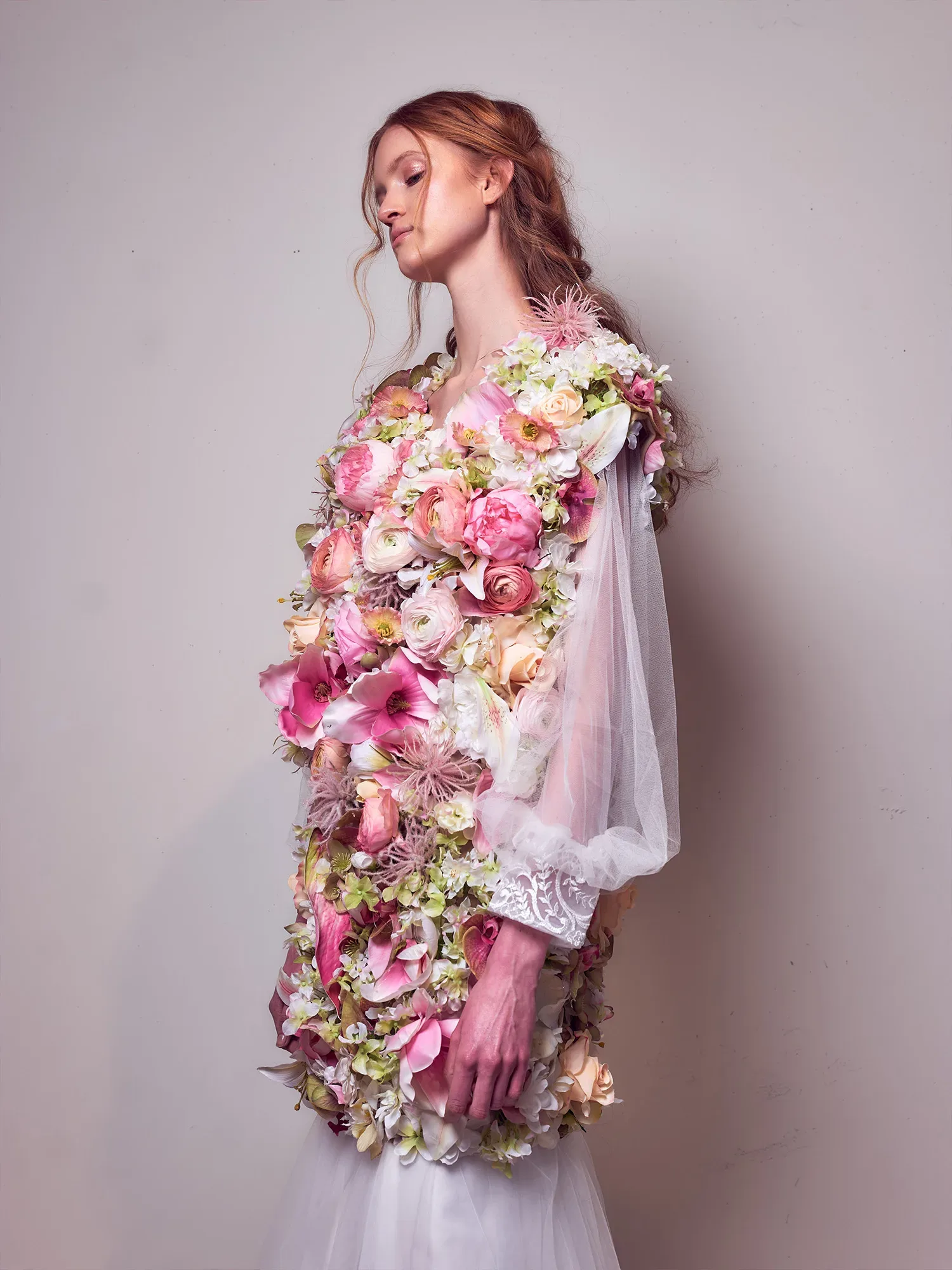Eine Frau trägt ein extravagantes Hochzeitskleid, das vollständig mit Ranunkeln und anderen Blumen in Rosa-, Creme- und Grüntönen verziert ist. Das Kleid hat transparente Ärmel und ein voluminöses Blumendesign, das ihre gesamte Silhouette bedeckt. Die Frau hat langes, rotes Haar und steht seitlich, ihr Blick ist nach unten gerichtet.