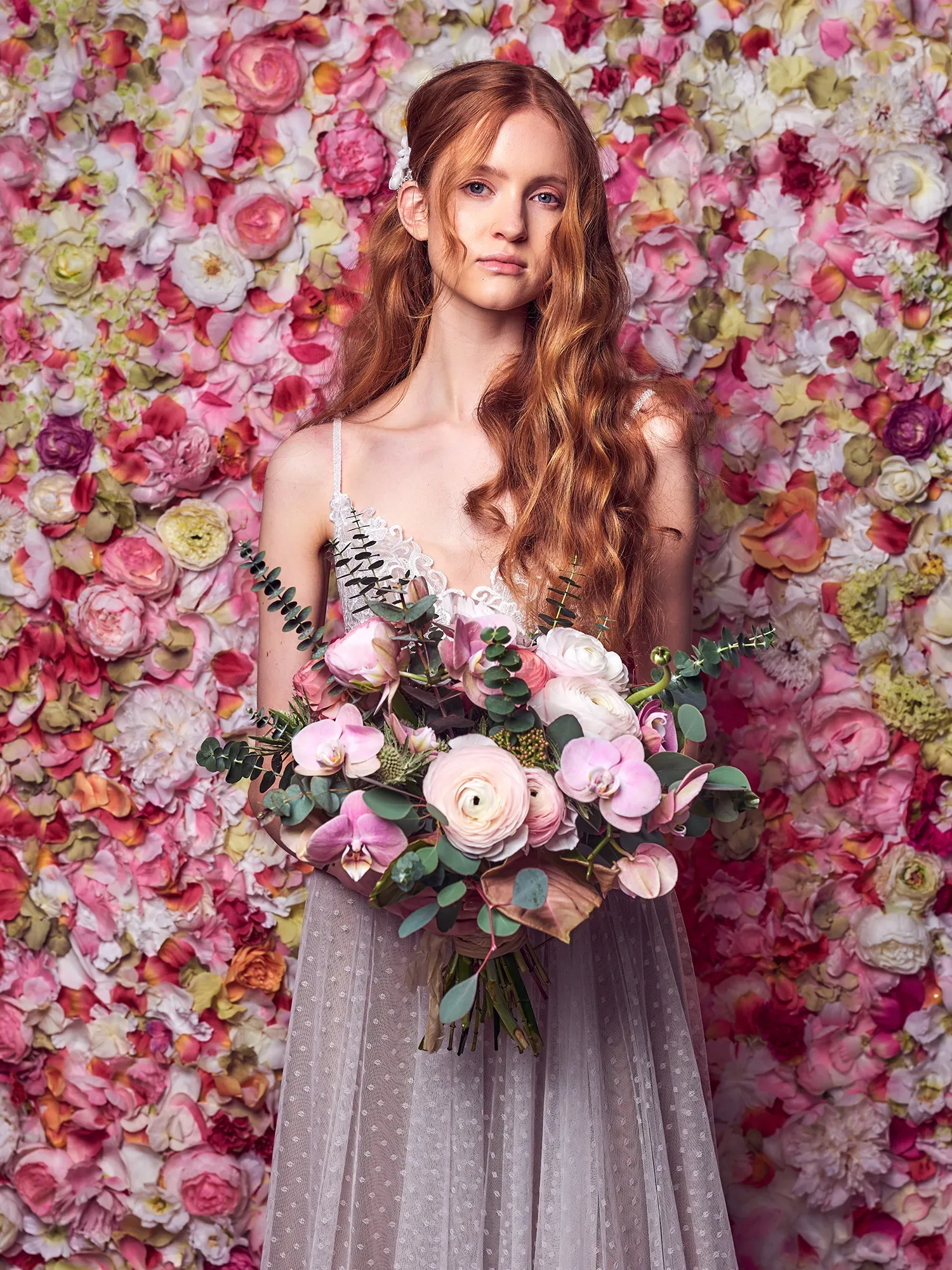 Eine Braut mit langem, rotem Haar steht vor einer üppigen Blumenwand aus Ranunkeln in verschiedenen Rosatönen und hält einen großen Strauß aus Ranunkeln, Orchideen und Eukalyptus. Ihr Kleid ist luftig und hat feine Spitzendetails, die ihr romantisches Aussehen ergänzen.