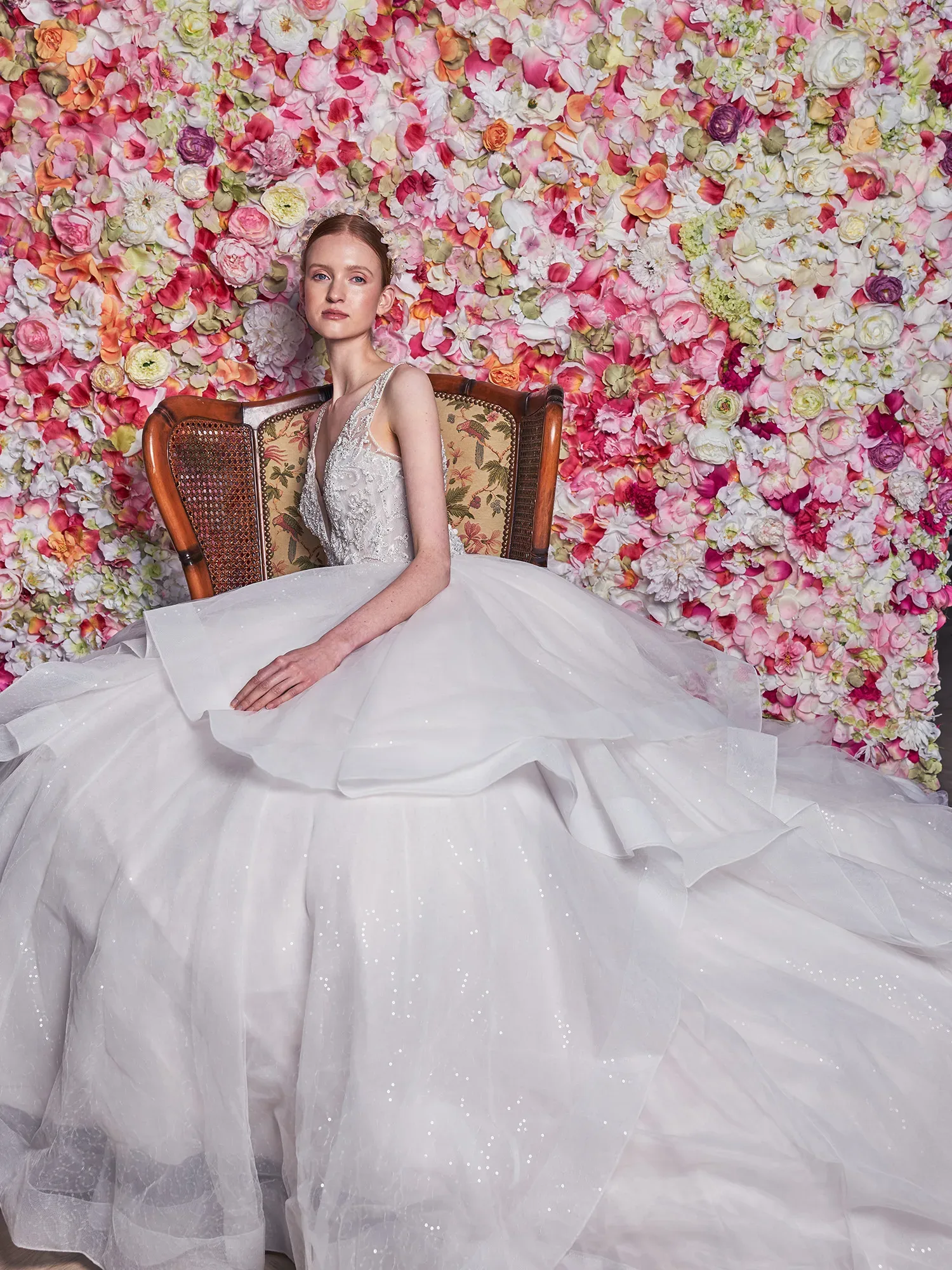 Eine Braut sitzt in einem prächtigen weißen Kleid auf einem antiken Stuhl vor einer Wand aus bunten Blumen. Das voluminöse Kleid aus mehreren Schichten Tüll fällt dramatisch zu Boden, während die Braut anmutig in die Kamera schaut. Die Blumenwand besteht hauptsächlich aus Ranunkeln in Rosa- und Cremetönen.