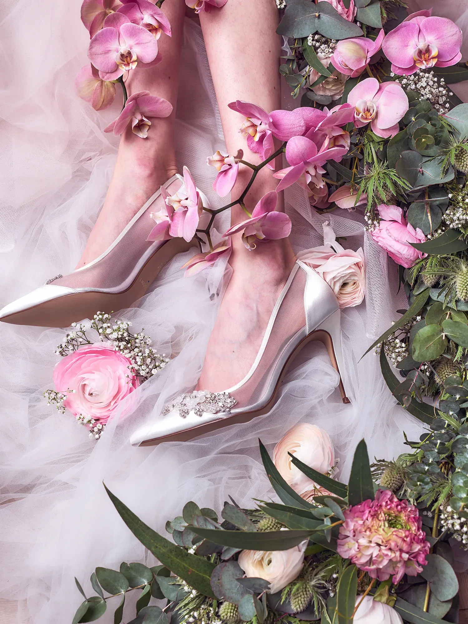 Eine Nahaufnahme von eleganten, spitzen Brautschuhen, die von rosa Orchideen und grünen Blättern umgeben sind. Die Brautschuhe stehen auf einem zarten, weißen Tüll, und neben ihnen sind rosa Ranunkeln und Eukalyptusblätter arrangiert, die einen romantischen und natürlichen Look erzeugen.