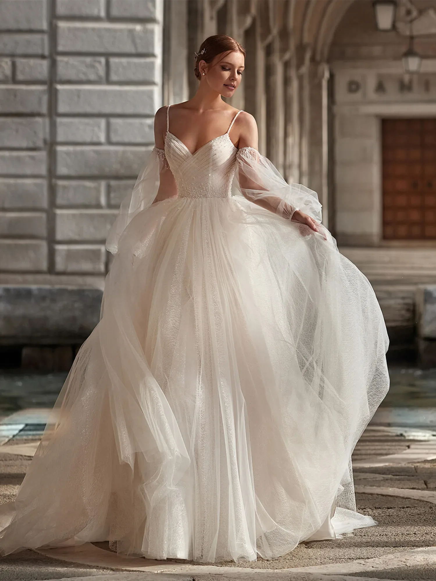 Eine Braut trägt ein trägerloses, romantisches Duchesse-Brautkleid aus Tüll mit abnehmbaren, transparenten Ärmeln. Sie steht vor einem historischen Gebäude und hebt leicht den Rock ihres Kleides, während der Wind sanft durch den Stoff weht.