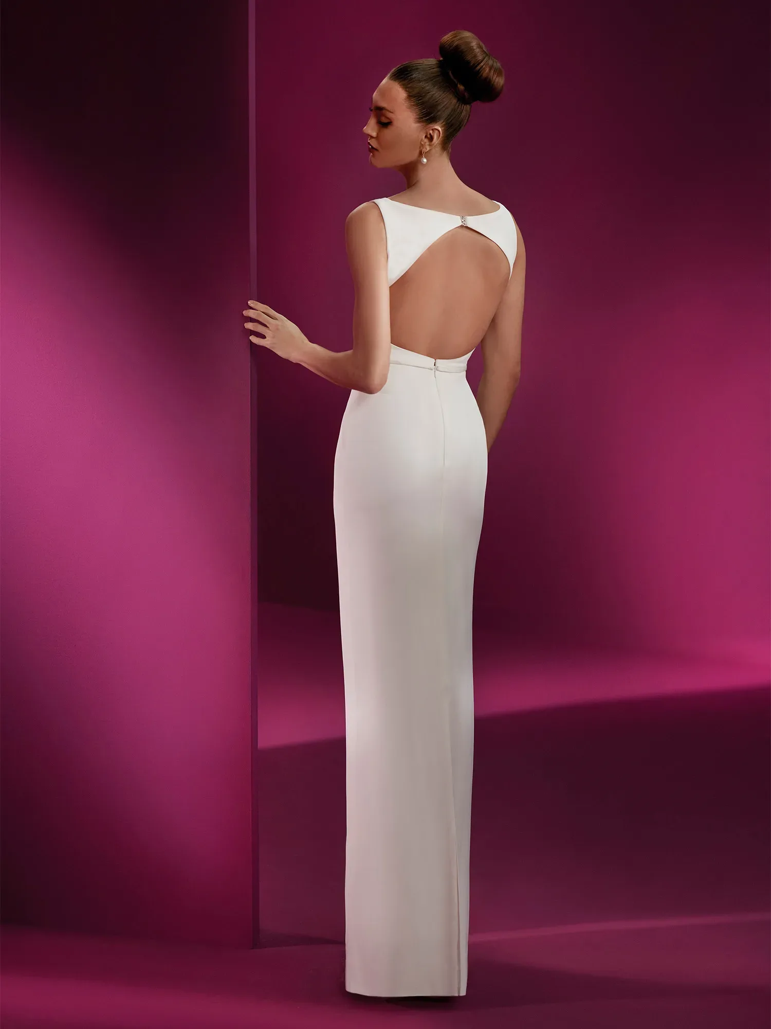 Eine Brautmodell in einem eleganten, figurbetonten Etui-Hochzeitskleid von Pronovias steht mit dem Rücken zur Kamera, die ein großzügiges Rückendekolleté zeigt. Das Kleid hat eine schlichte und raffinierte Ästhetik mit einem minimalistischen Design, das durch einen kleinen Diamantenverschluss am Rücken akzentuiert wird. Ihr Haar ist zu einem kunstvollen Knoten gestylt. Sie steht vor einem leuchtend pinken Hintergrund.