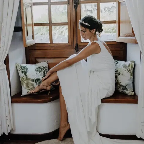 Braut sitzt auf einer Bank unter einem Fenster und zieht sich ihr Schuhe an