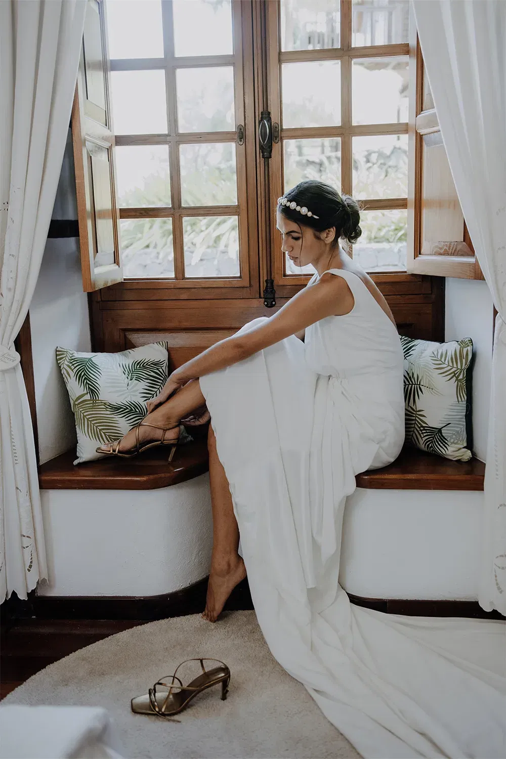 Braut sitzt auf einer Bank unter einem Fenster und zieht sich ihr Schuhe an