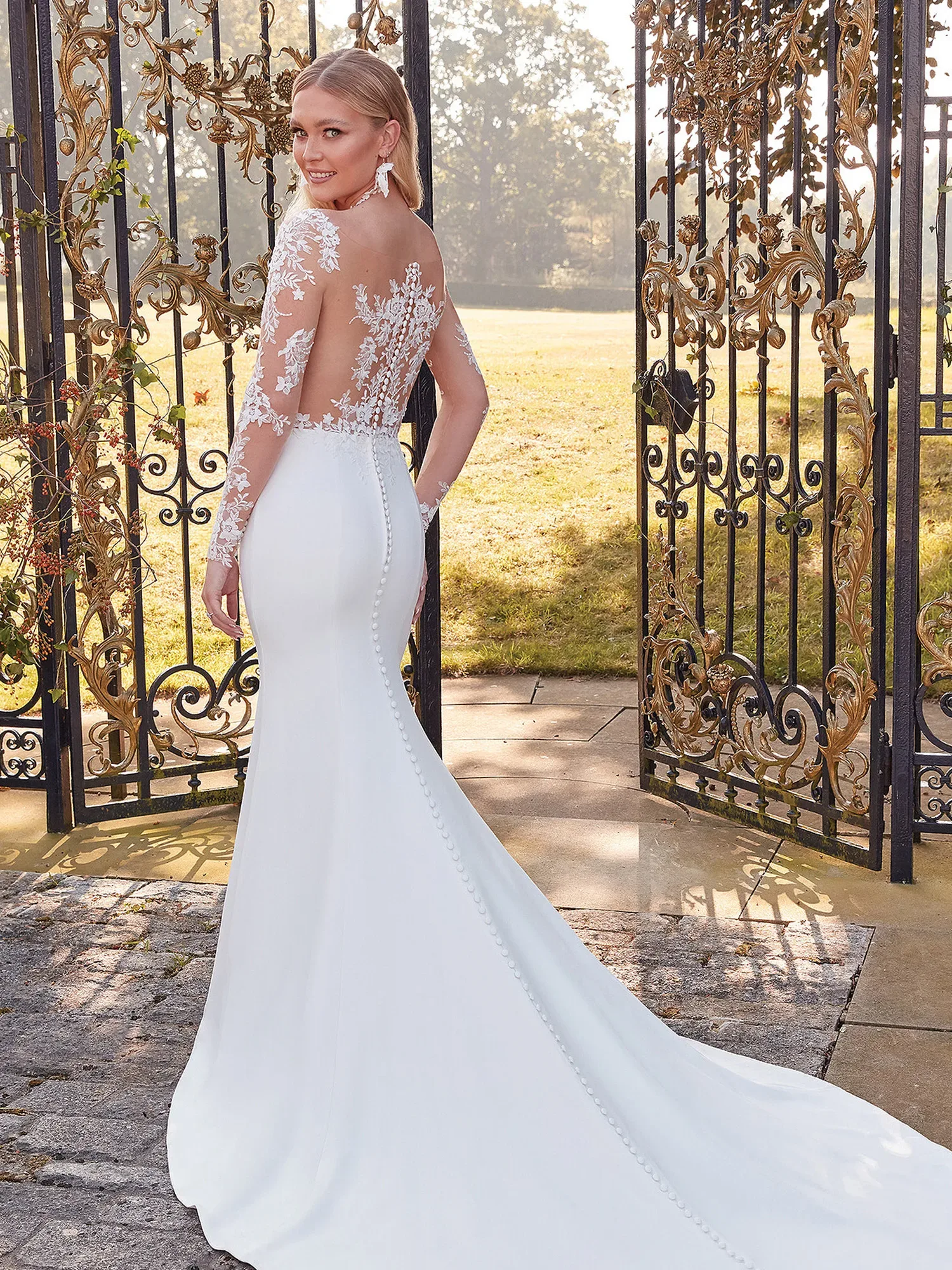 Eine Braut in einem eleganten Godet-Brautkleid mit langen Ärmeln aus Spitze und einem tiefen Rückenausschnitt steht vor einem kunstvoll verzierten Gartentor. Der bodenlange Rock betont ihre Silhouette, während sie über die Schulter lächelt.