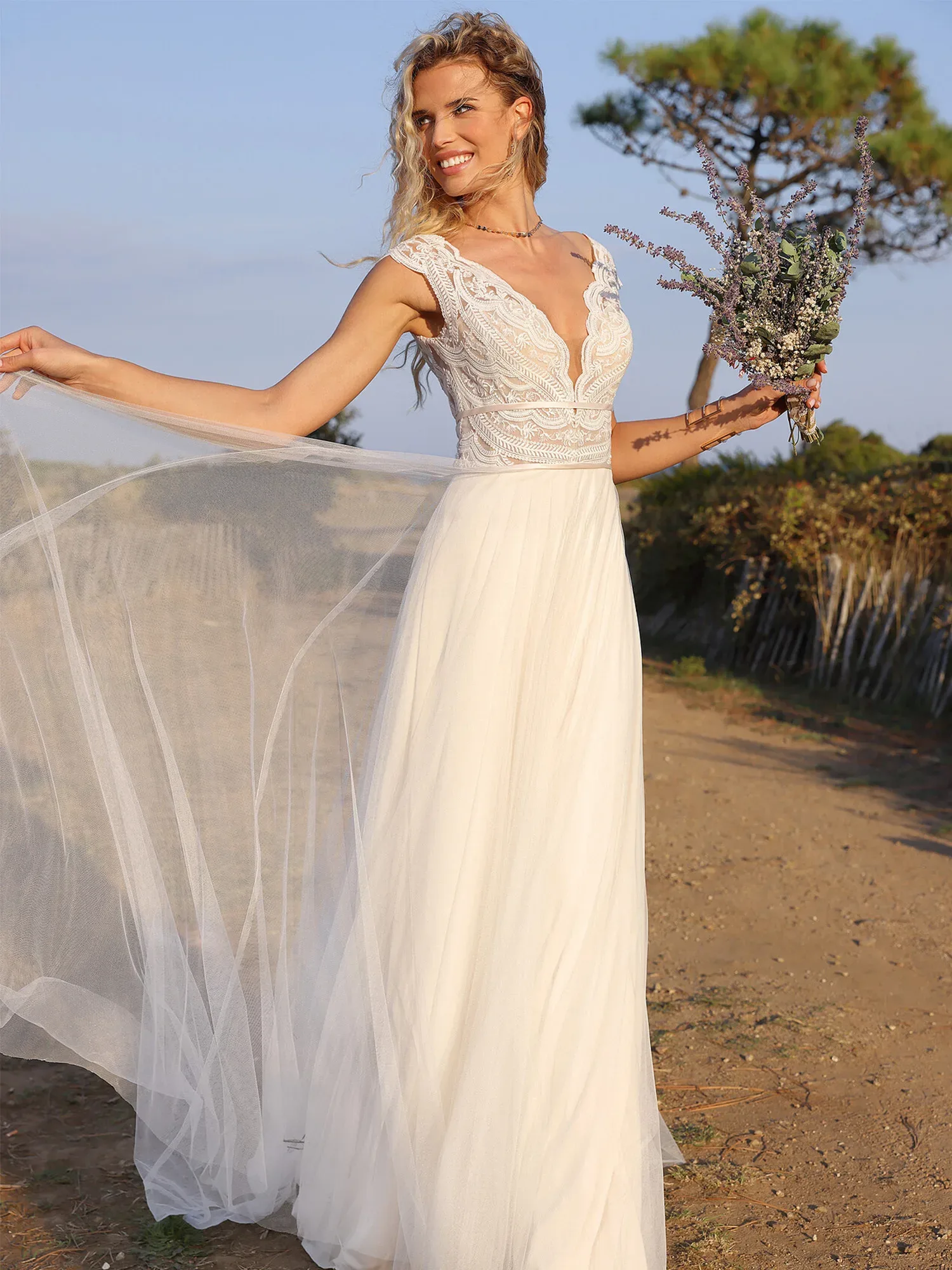 Eine Braut steht in einem weißen Hochzeitskleid im i-Linien-Stil, mit einem Brautstrauß aus Lavendel und anderen grünen Zweigen in der Hand, auf einem erdigen Weg, der von einen Holzzaun und Büschen geführt wird. Das Kleid hat ein Spitzenoberteil mit tiefem V-Ausschnitt und geht in einen fließenden Tüllrock mit mehreren Schichten über..
