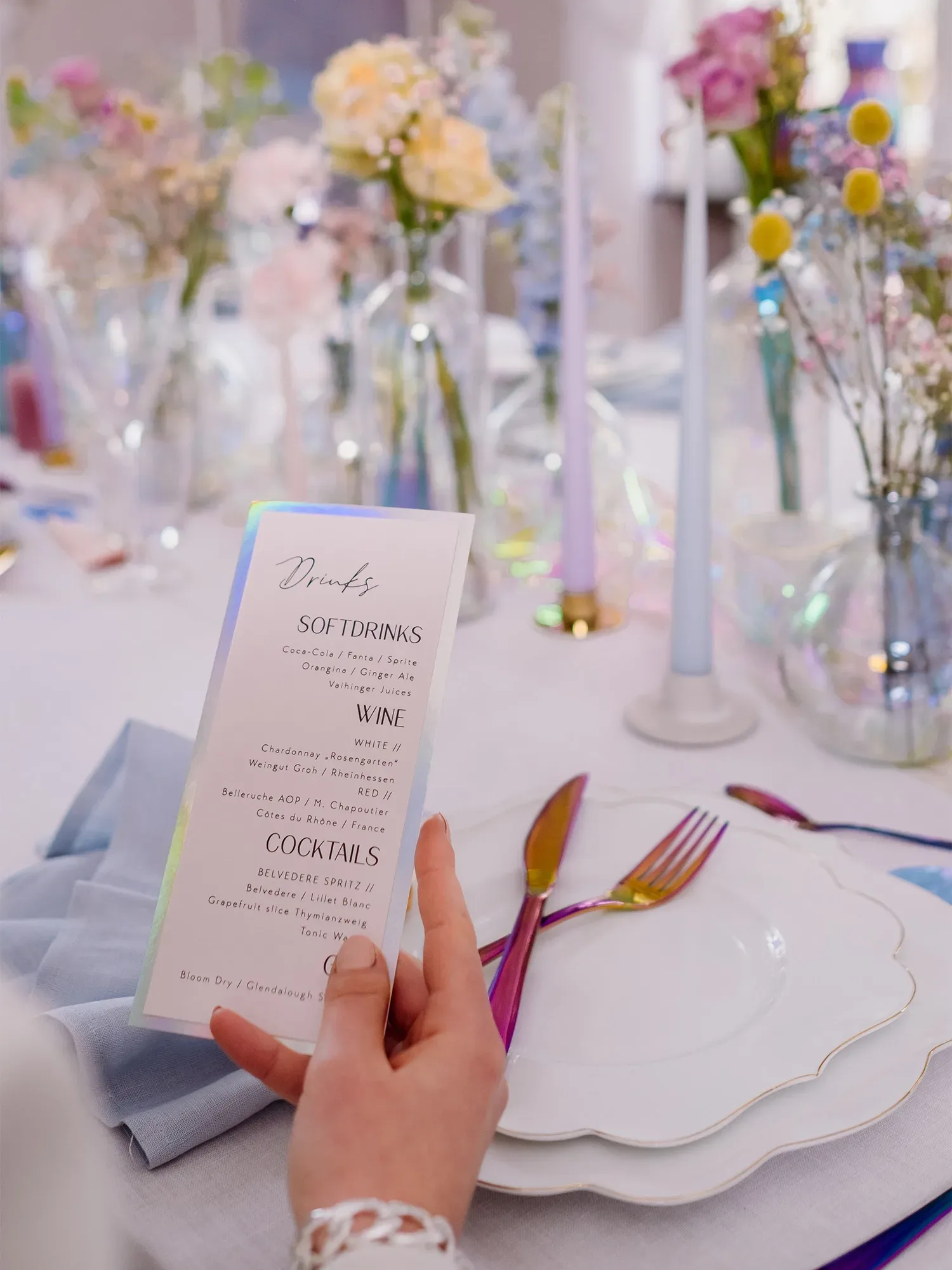Nahaufnahme einer Hand, die eine Getränkekarte an einem stilvoll gedeckten Tisch hält. Auf dem Tisch sind bunte Blumenarrangements in verschiedenen Glasvasen verteilt, begleitet von pastellfarbenen Kerzen. Das Besteck ist in metallischen Regenbogenfarben gehalten, und die Teller haben feine, goldene Ränder.