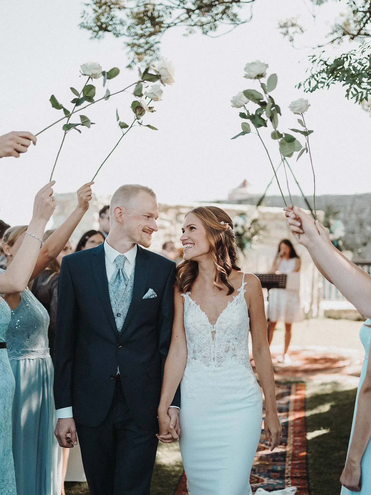 Das Brautpaar läuft lächelnd den Gang entlang, während Freunde und Familie sie mit Rosenstängeln in die Ehe geleiten. Die Braut trägt ein elegantes, weißes Spitzenkleid, und der Bräutigam ist in einem dunkelblauen Anzug gekleidet. Die Hochzeitsgesellschaft steht jubelnd um sie herum.