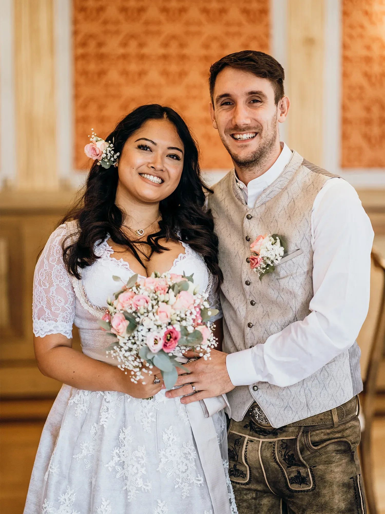 Ein Brautpaar lächelt glücklich in die Kamera. Die Braut trägt ein traditionelles, weißes Spitzenkleid mit floralen Akzenten und hält einen Blumenstrauß in Rosa und Weiß. Der Bräutigam trägt eine beige Trachtenweste und Lederhosen.