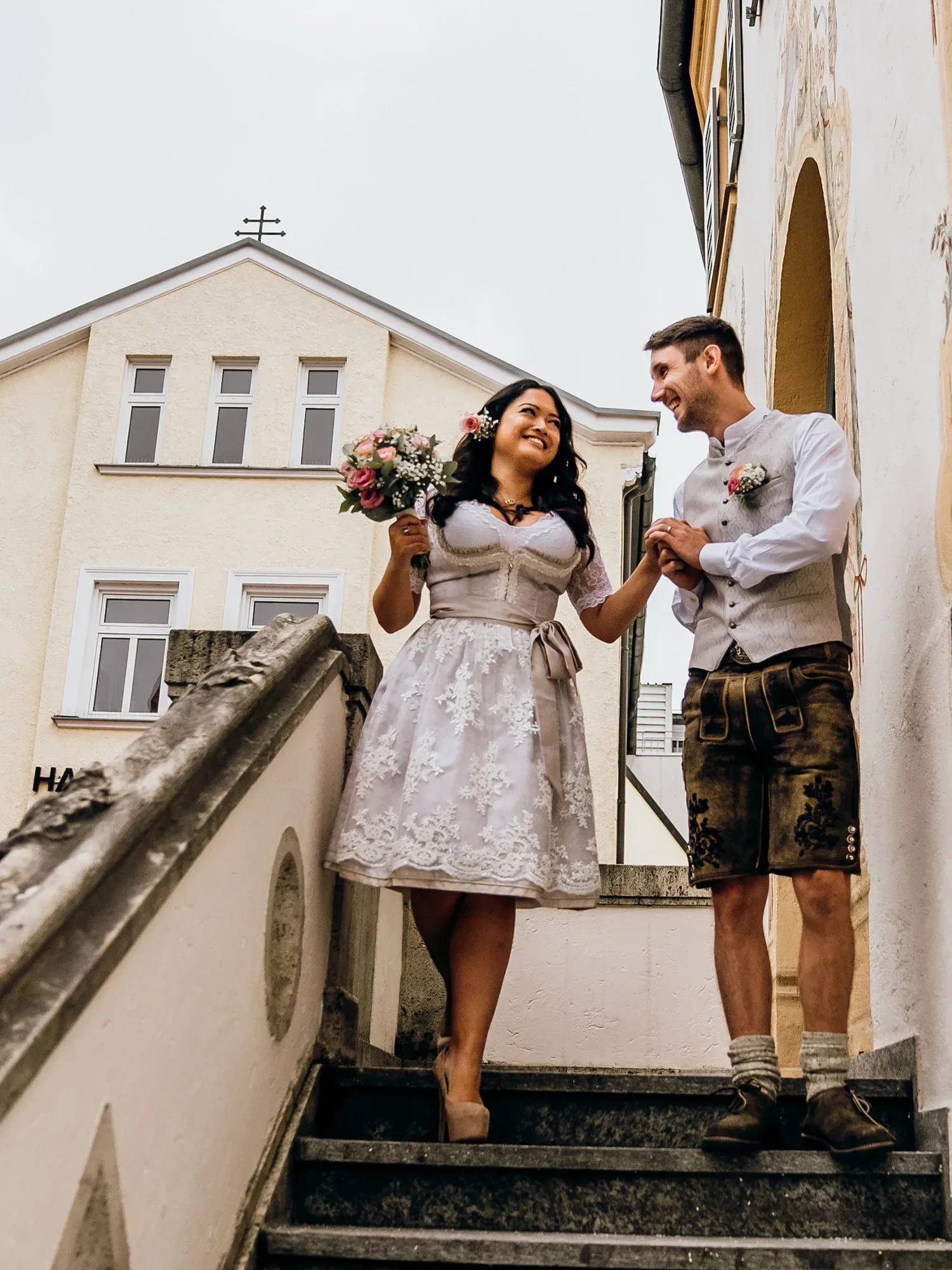 Die Braut und der Bräutigam gehen eine Treppe hinunter, während sie sich an den Händen halten und einander liebevoll ansehen. Die Braut trägt ein weißes Dirndl, und der Bräutigam trägt eine Lederhose mit Trachtenhemd. Im Hintergrund ist ein traditionelles Gebäude zu sehen.