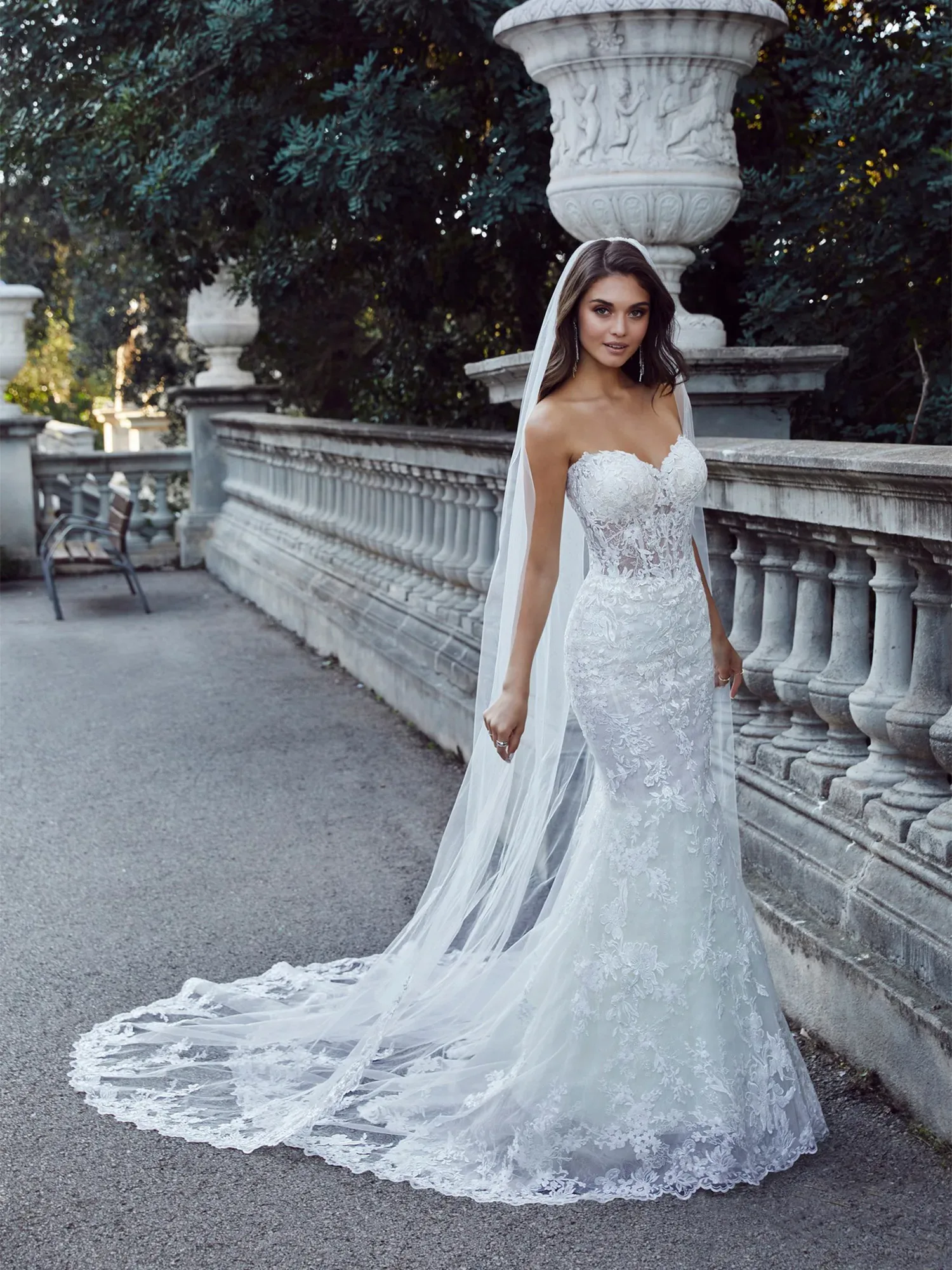 Eine Braut trägt ein figurbetontes Mermaid-Brautkleid mit floralen Stickereien und einer langen, transparenten Schleppe. Sie steht an einem historischen Gebäude und trägt einen langen Schleier, der den eleganten Look unterstreicht.