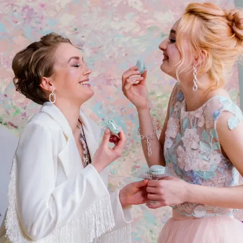 Zwei Frauen stehen fröhlich beieinander und lachen, während sie sich gegenseitig Macarons reichen. Eine Frau trägt einen weißen Blazer mit Fransen, die andere ein elegantes Oberteil aus Spitze in Pastelltönen. Der Hintergrund ist eine bunte, abstrakte Wand.